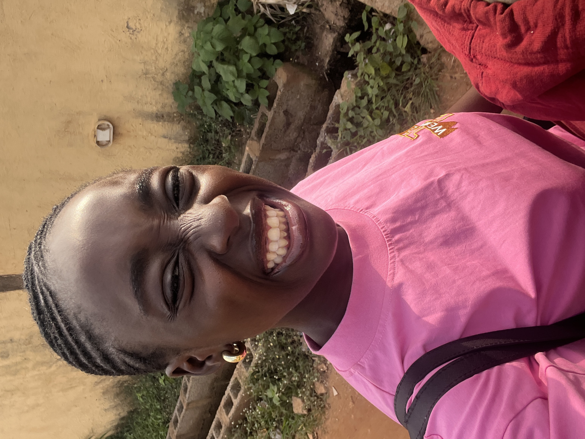 In Galadimawa, Nigeria, a young woman with braided hair and a gold hoop earring in her left ear is captured in a close-up, outdoor, eye-level shot. She is wearing a bright pink t-shirt with a graphic design that includes the word "WE." Her face is contorted in a wide, toothy grin, with her eyes squinted and the skin around them wrinkled, suggesting laughter or an intense expression. Her teeth are visibly white. She is carrying a black bag strap over her left shoulder, and a portion of a red garment is visible on her right. The background consists of a textured yellow wall, some greenery, and a rough, uneven ground with what appears to be a ditch or drainage area. The lighting suggests it is daytime, possibly late afternoon, with shadows casting from the right.