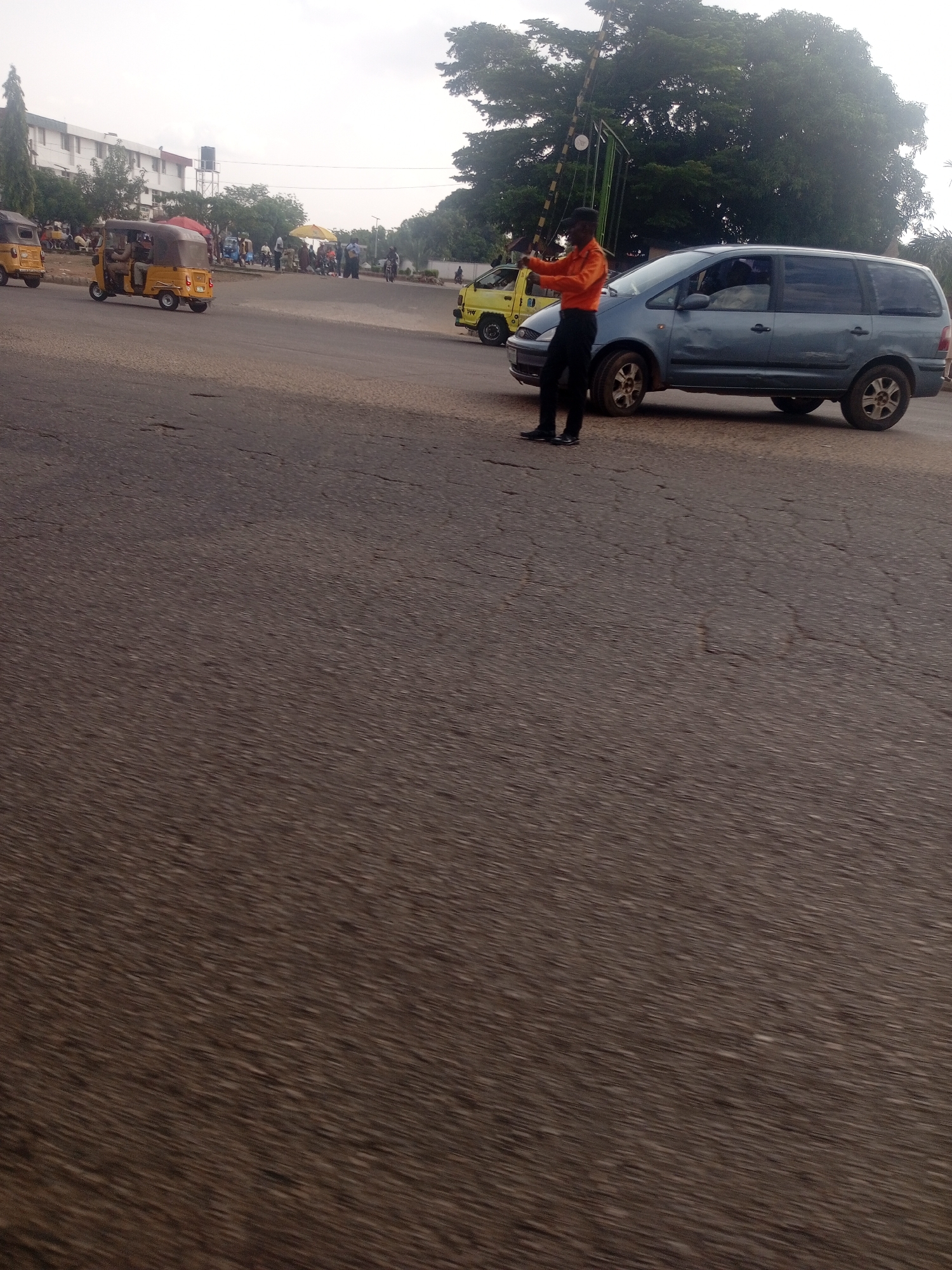 The image depicts a street scene in Unguwan Dosa, Nigeria. The foreground is dominated by a textured, asphalt road, suggesting it's a busy thoroughfare. In the midground, a man in an orange shirt and black trousers stands directing traffic. He appears to be a traffic controller or police officer. He is positioned next to a light blue minivan, which is stopped. Behind the minivan, a yellow car is also visible.
To the left of the man and the minivan, a yellow auto-rickshaw, also known as a "Keke Napep," is moving. Several other auto-rickshaws and people are present in the background, clustered around what appears to be a market or waiting area near some buildings. The buildings are light-colored and have multiple windows. A tall tree with dense foliage stands behind the traffic controller and minivan. There are also some market stalls with yellow umbrellas visible in the distance. The sky is overcast, indicating it might be a cloudy day or early morning/late afternoon. The overall impression is of a lively, functional urban environment. There is no visible text in the image.