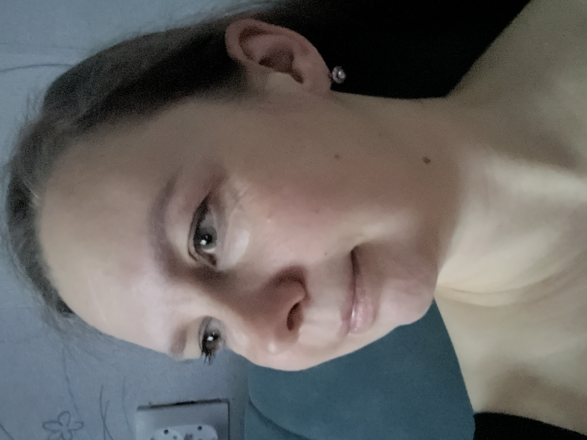 A close-up, angled portrait features a person, likely female, with fair skin, dark eyes, and dark hair. The image is rotated counter-clockwise, showing the subject's head tilted, looking downwards with a soft, gentle smile. Her dark hair is pulled back, revealing the side of her face and an ear adorned with a small, circular earring. She is wearing a dark top, possibly black or dark blue, visible on her shoulder and upper chest.
The background is softly blurred. On the left, a light blue-gray wall is visible, featuring a faint, lighter pattern resembling a flower or abstract shape, and a white electrical outlet with two circular sockets. To the right, a darker, possibly teal or deep blue, surface suggests another part of the indoor setting, perhaps furniture or a curtain. The lighting is soft and even, consistent with an indoor environment. There is no visible text. The image captures a quiet, personal moment, possibly a selfie, within an indoor location in Chaikovsky, Russia.