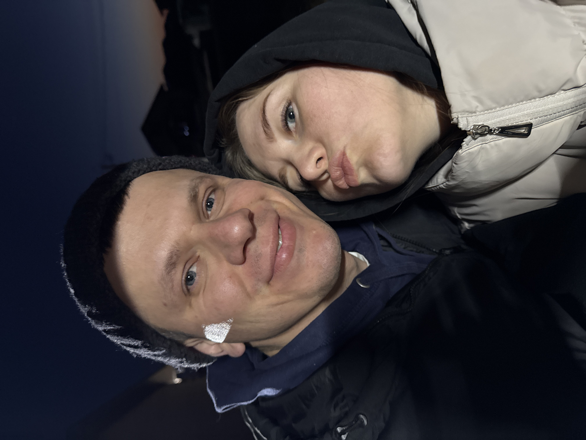 The image shows a selfie of two people. A man with short hair and a black beanie is on the left, smiling at the camera. He has a shiny, heart-shaped object stuck to his cheek. Next to him, a woman with blonde hair, wearing a black hoodie and beige winter coat, is kissing the air. The background is dark. The location is Gornyi (Dvurechenskoe S/P), Russia.