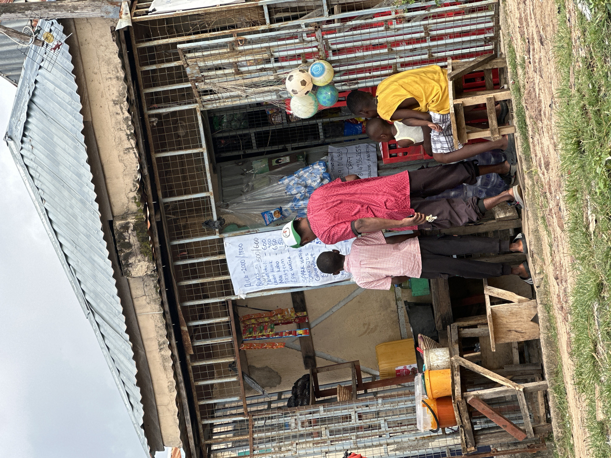 A street vendor stall in Narayi, Nigeria, is the setting for this image. The stall is constructed with corrugated metal roofing and a barred storefront, suggesting security measures. A whiteboard displaying prices for various food items, including rice, beans, and vegetables, is prominently displayed. Several people are visible, including two young boys and an adult male wearing a red shirt, who appear to be customers or associates of the vendor. One of the boys is seated on a wooden stool, while the other stands with the man. The stall is stocked with goods such as drinks, bags of snacks, and plastic containers filled with what appears to be food products. The environment is slightly overcast, and the ground is covered in a mixture of dirt and grass. The overall scene depicts a typical urban marketplace in Nigeria.