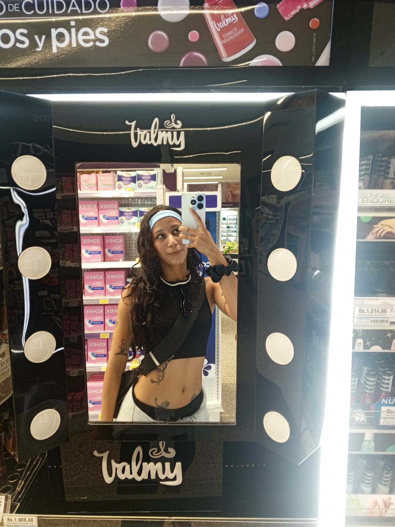 The image shows a woman taking a selfie in a retail environment, presumably a drugstore or supermarket in Pampatar, Venezuela. She is standing in front of a "Valmy" branded mirror-like display, which is illuminated by circular lights. The display is flanked by shelves stocked with products, including feminine hygiene items and cosmetics. The woman is wearing a black crop top, light-colored pants, and a blue headband. She has dark curly hair and tattoos on her torso. The shelves behind her contain products like Kotex pads and Valmy nail polish. Price tags in Venezuelan bolivars are visible on the shelves adjacent to the display. The overall setting suggests a modern retail space within the city of Pampatar.
