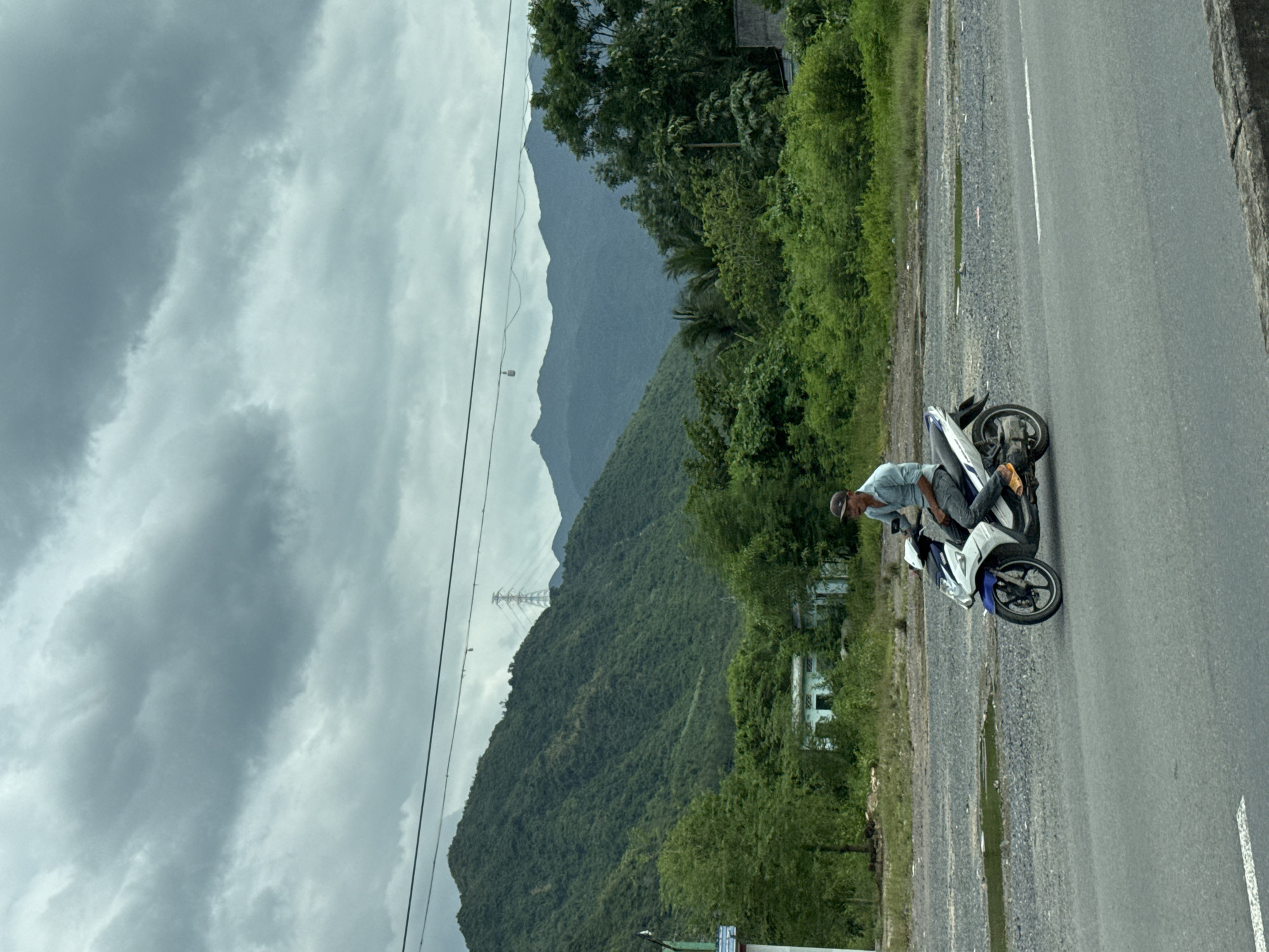 A man in a light blue button-down shirt and dark pants rides a white and blue scooter along a paved road. He is wearing a dark baseball cap and yellow sandals. The scooter has a white front fairing with blue accents and a black rear fender. The road has a white lane marking. In the background, a heavily vegetated mountain range under a cloudy sky dominates the scene. There are power lines strung across the upper part of the image, with a pylon visible on the mountainside. A small, light-colored building with visible windows is situated amidst the greenery in the mid-ground. The location is the outskirts of Nam Ninh Hòa, Vietnam.