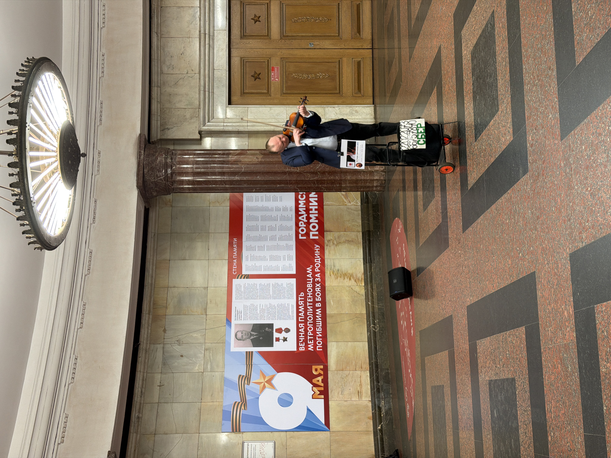 The scene depicts a man playing a violin in the Moscow Metro, Russia. He stands in front of a commemorative poster titled "WALL OF MEMORY" in Russian and dedicated to the metro workers who perished in battles for their homeland, with the "9 MAY" (Victory Day) date prominently displayed. The man is dressed in a dark suit jacket and a light-colored shirt, holding and playing his violin. Next to him is a small trolley with a sign on it, possibly indicating his name or the music he plays. A speaker system is positioned nearby, likely used to amplify the sound of his violin. The Metro station features a large, ornate chandelier and marble walls, with a distinctive geometric pattern on the floor. A wooden door with star decorations on it can be seen in the background.