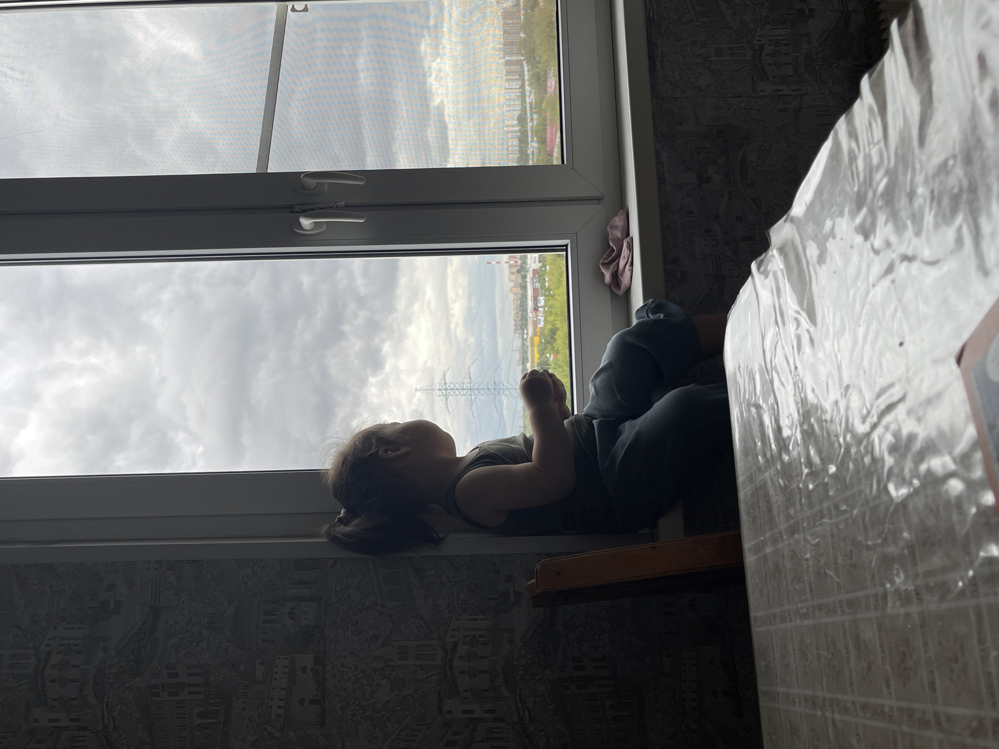 A young child with dark hair tied in a ponytail sits on a windowsill, facing away from the camera and looking out of a large window. The child is wearing a dark sleeveless shirt and dark pants. Next to the child on the windowsill is a pink folded cloth. The window overlooks a city landscape with buildings, trees, and a power line tower under a cloudy sky. To the left of the child, a wooden chair is visible, and behind the child, the wall is covered with patterned wallpaper depicting buildings. In the foreground, the edge of a table covered with a transparent plastic sheet is visible.