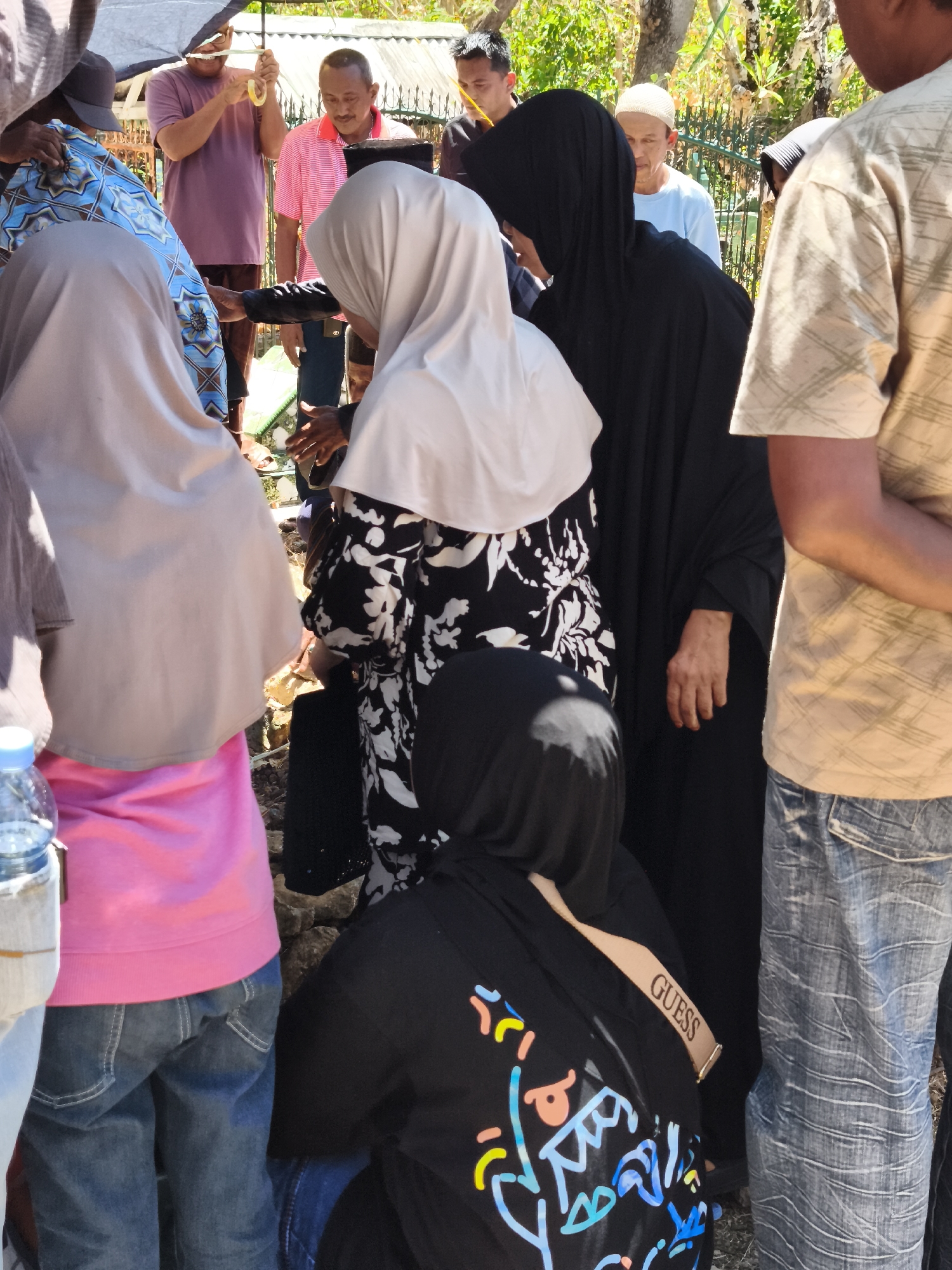 The image depicts a gathering of people outdoors in what appears to be a village or cemetery in Tonasa, Indonesia. The scene is characterized by a mix of formal and casual attire, suggesting a significant event, possibly a funeral. Several individuals are wearing traditional Islamic clothing, including hijabs and kufis, while others are in everyday wear like t-shirts and jeans.
In the foreground, two women are partially visible. One is wearing a pink long-sleeved shirt and blue jeans, her back to the camera. The other is seated, wearing a black hoodie with a colorful graphic design and a beige "GUESS" logo strap across her shoulder.
Behind them, several individuals are interacting. A woman in a black hijab and abaya is standing, her hand visible. Next to her, a woman in a light-colored hijab and a black and white floral dress is also present. Further back, a man in a pink and white striped shirt is holding an umbrella with another person. Another man in a purple t-shirt and dark pants is also visible, along with others in the background.
The environment suggests a sunny day with lush greenery in the background, trees, and what appears to be a low metal fence. The ground is uneven and rocky. A blue plastic water bottle is also visible in the lower left corner. The overall impression is one of a communal gathering, likely a solemn occasion given the attire and the presence of what could be a grave site.