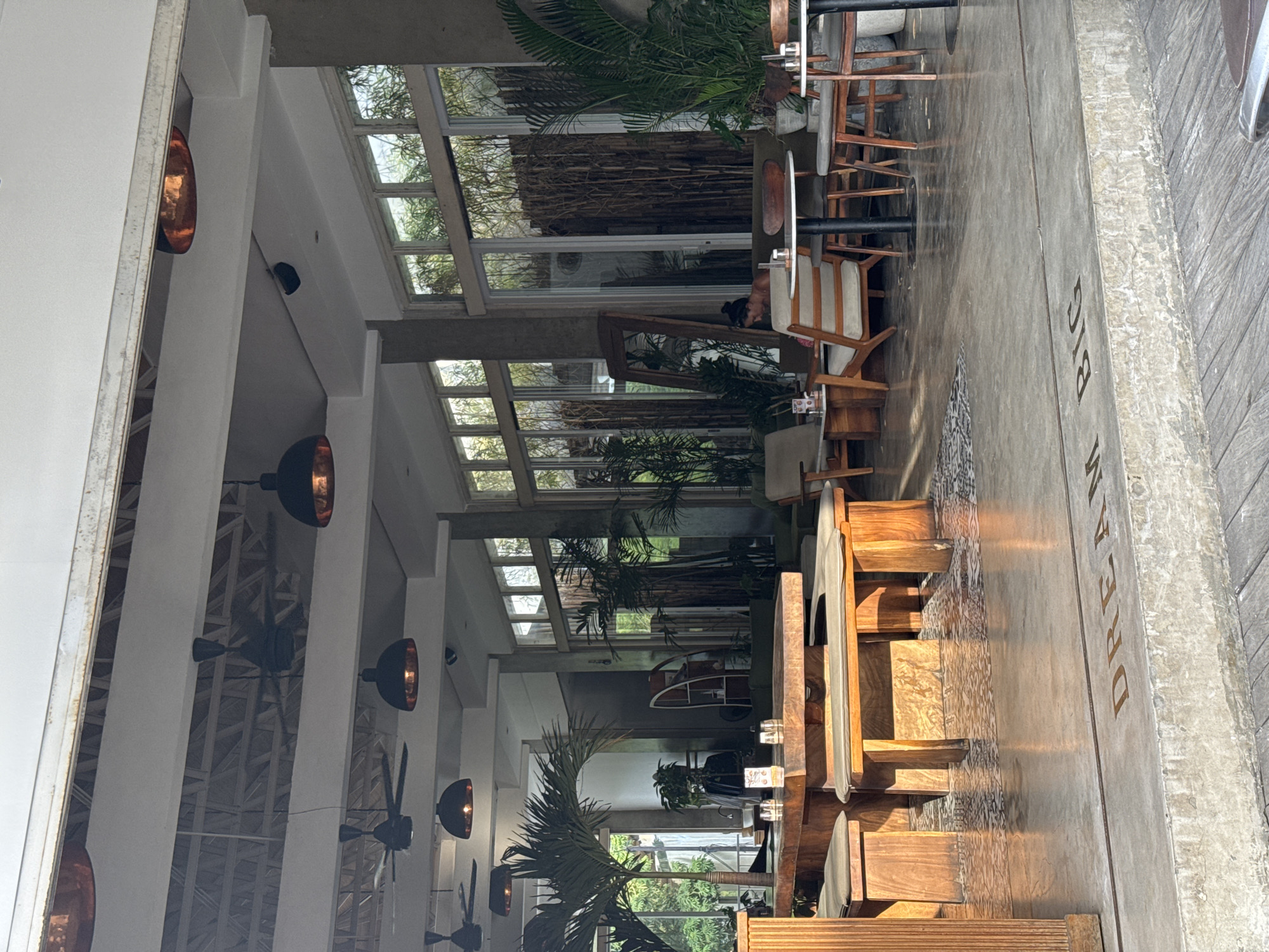 The image shows the interior of a restaurant in Kuta Selatan, Indonesia, during daytime. The space is characterized by a modern, open design with a high ceiling featuring numerous windows and supporting pillars. Rows of copper-colored pendant lights and ceiling fans are visible along the ceiling. Lush green plants and palm trees are placed throughout the restaurant, giving it a tropical ambiance.
Wooden tables and chairs are arranged for dining, with some seating areas partially visible near the entrance. A woman is seated at one of the tables near the back of the restaurant. A sign with the words "DREAM BIG" is visible on the floor near the entrance. The image suggests a relaxed, upscale dining environment in a tropical setting.