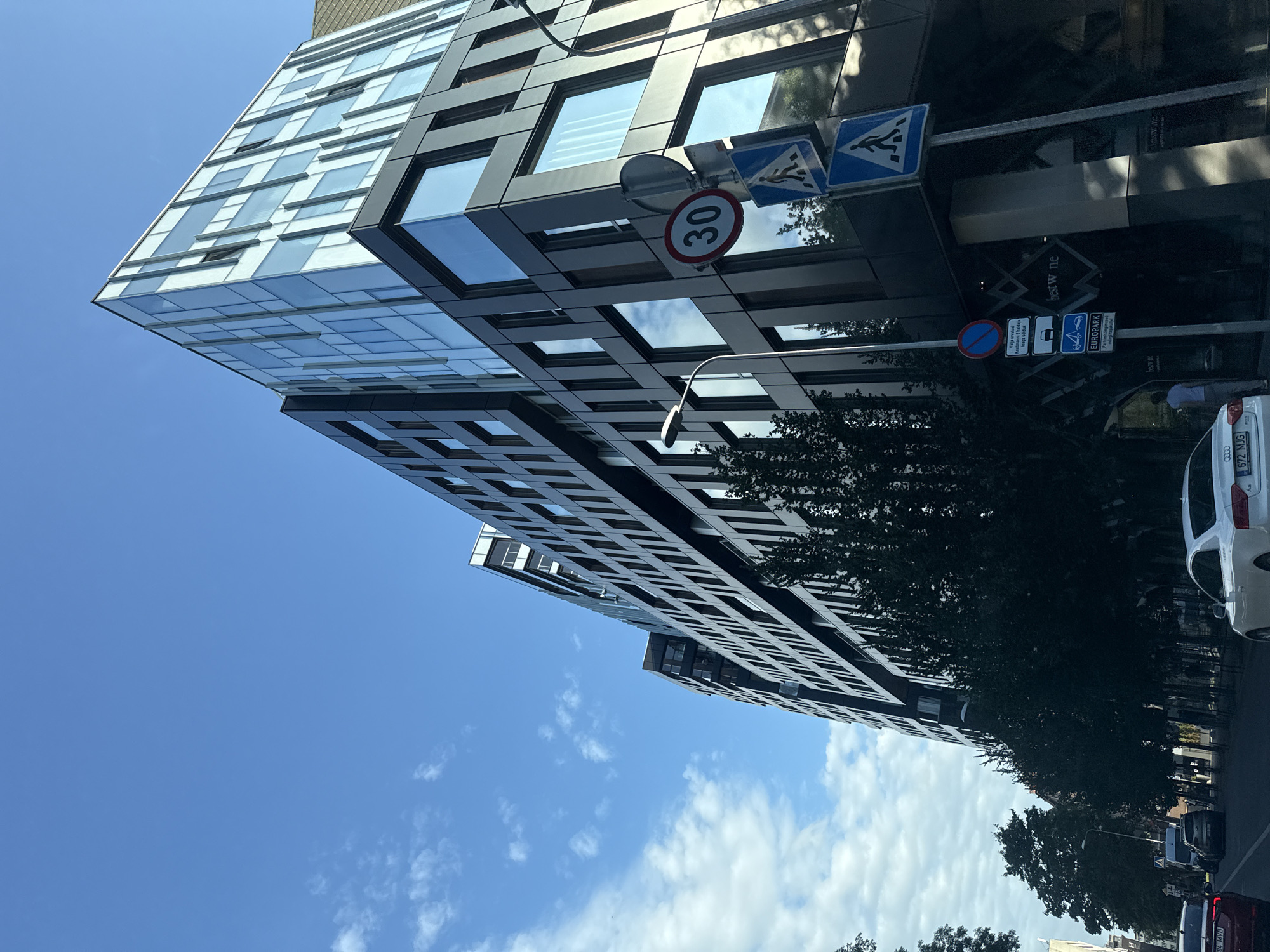 The image captures a contemporary building on a street in Tallinn, Estonia. The building features a modern architectural design with dark metallic panels and extensive glass facades, reflecting the bright blue sky and scattered white clouds. Several traffic signs are visible, including a speed limit sign indicating "30," and pedestrian crossing signs. A street lamp stands next to the building. Trees line the street in front of the building. Several cars are present on the street, including a white Audi sedan in the foreground. The overall scene suggests a typical urban environment during the daytime.