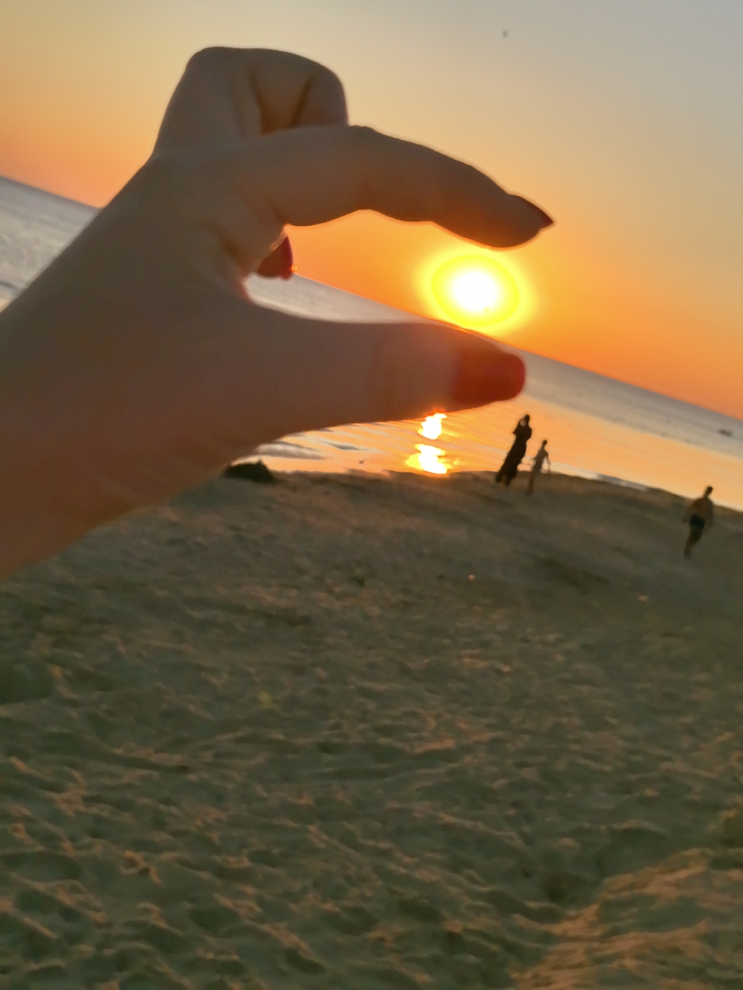 A person's hand, with red painted fingernails, is positioned in the foreground, appearing to hold the setting sun. The sun is a bright, golden orb, casting a warm glow across the sky and reflecting on the water. The scene takes place on a sandy beach, with the sea stretching out to the horizon. In the background, several silhouetted figures of people are visible walking or standing on the beach. The overall atmosphere is peaceful and serene, characteristic of a sunset on a beach, possibly in Anapa, Russia, a city known for its Black Sea coastline and popular tourist destinations.