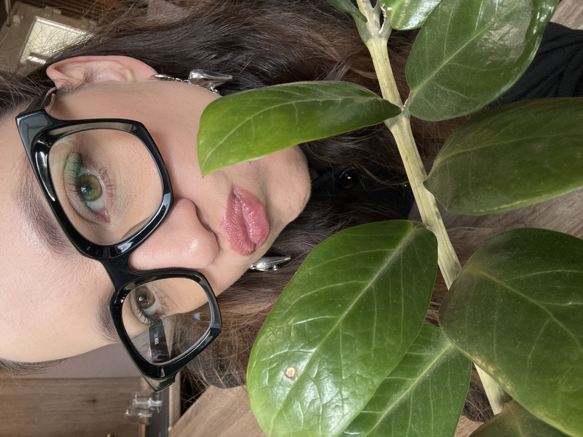 A close-up selfie shows a woman wearing large black-rimmed glasses and silver earrings with wing-shaped charms. She is puckering her lips, which are covered in pink lipstick. Her eyes are green and her eyebrows are dark. Green leaves from a plant are in the foreground, obscuring part of her face and the background. The background appears to be indoors with some wooden surfaces and what might be glasses on a shelf. The image does not provide any information about the city of Chaikovsky, Russia.