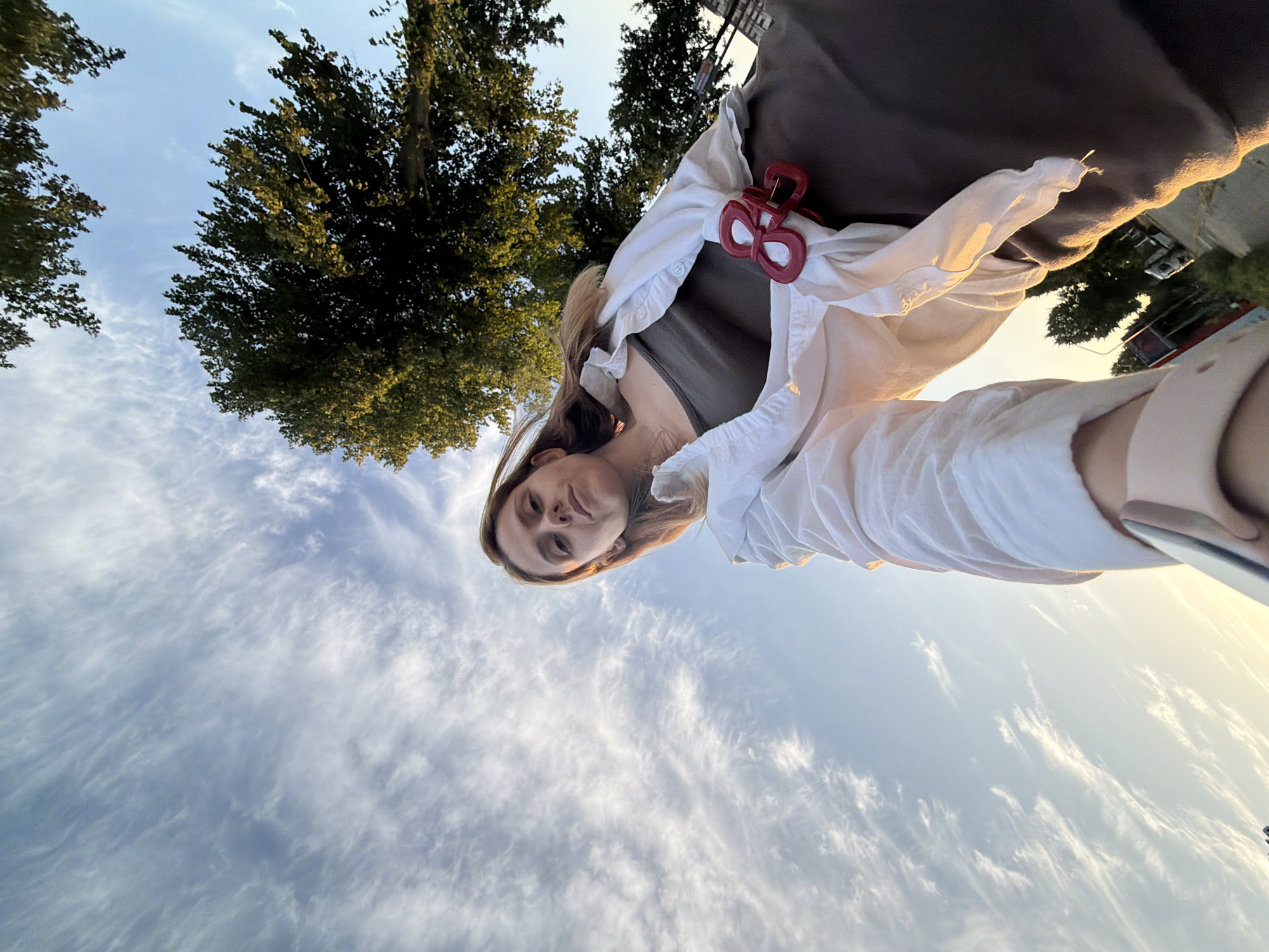 A low-angle, wide-shot, outdoor photograph captures a young woman standing below tall trees in Helmond, Netherlands. The woman, with light brown hair and a serious expression, is wearing a dark tank top, a white unbuttoned shirt, and a brown skirt. A red hair clip is attached to her shirt. Her left arm is extended towards the camera, holding a phone in a selfie pose. The sky is blue with wispy white clouds, and sunlight illuminates the scene, casting long shadows. In the background, greenery from the trees and a street with a white van are visible.