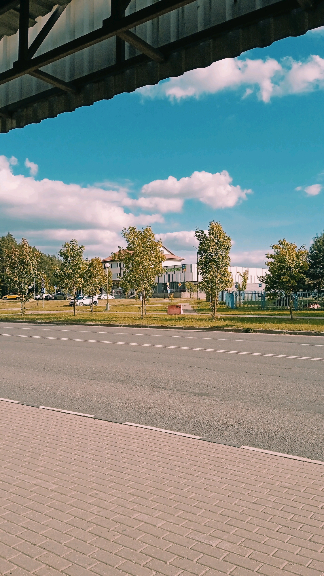 The image shows a street scene in Бараўлянскі сельскі Савет, Belarus, under a bright blue sky with scattered white clouds. A paved sidewalk runs along the bottom of the frame, transitioning to an asphalt road with white lane markings. Across the road, a grassy median is lined with young deciduous trees. In the background, a two-story building with a light-colored facade and a darker roof is visible, with signage in Cyrillic that reads "СУПЕРМАРКЕТ". A blue metal fence runs along the property line. Several cars are parked in front of the building and on the street. The overall impression is of a suburban or commercial area on a clear day.