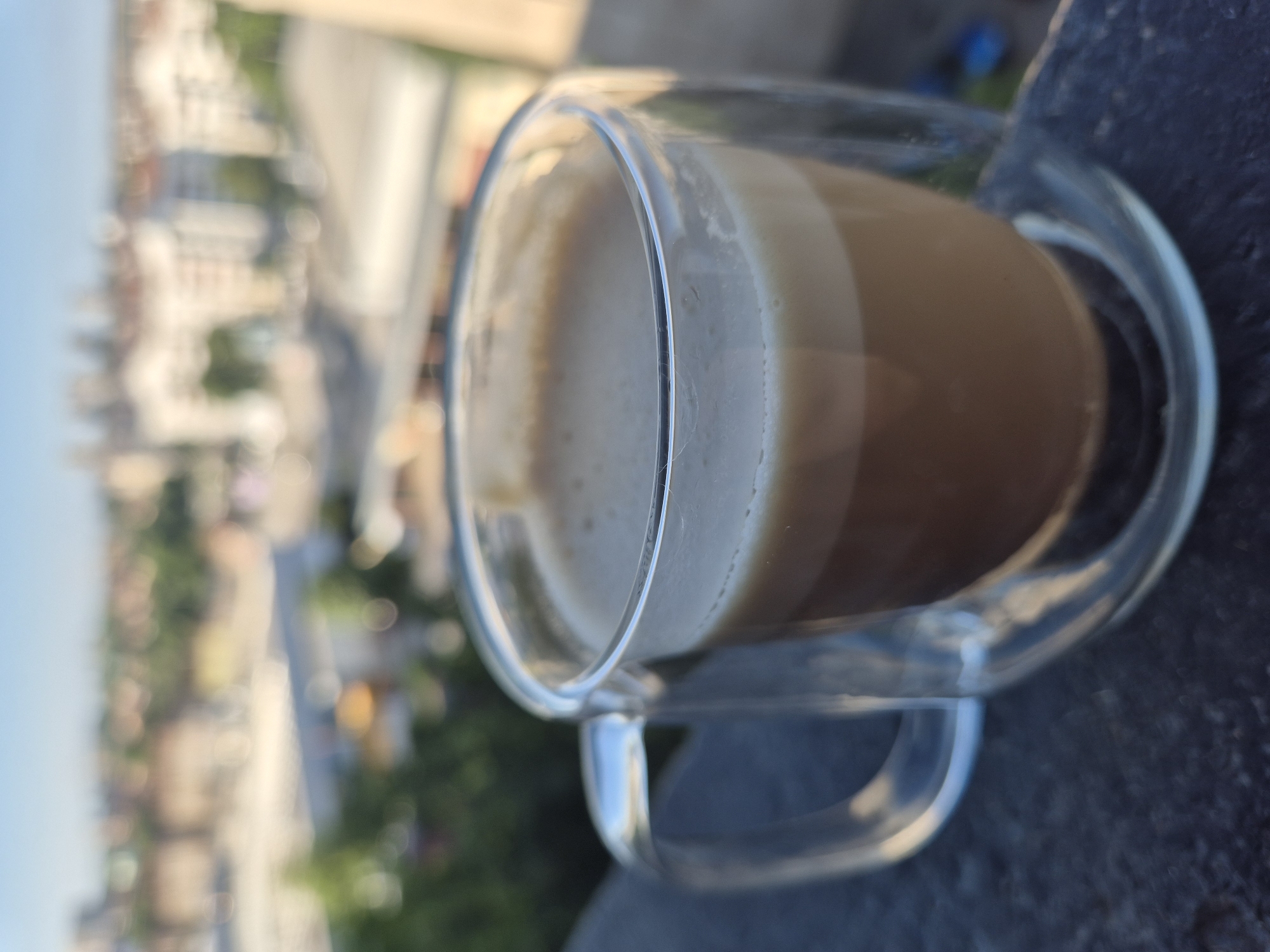 A double-walled glass mug of coffee with foam sits on a dark, textured surface, likely a balcony railing, with a blurred cityscape in the background under a clear blue sky. The mug is made of clear glass, revealing the layered coffee within, with a distinct layer of light brown foam on top. The handle of the mug is also made of clear glass and curves outwards. The surface the mug is resting on is dark and appears to be made of stone or concrete. The background shows an out-of-focus view of buildings and trees, suggesting an urban environment.