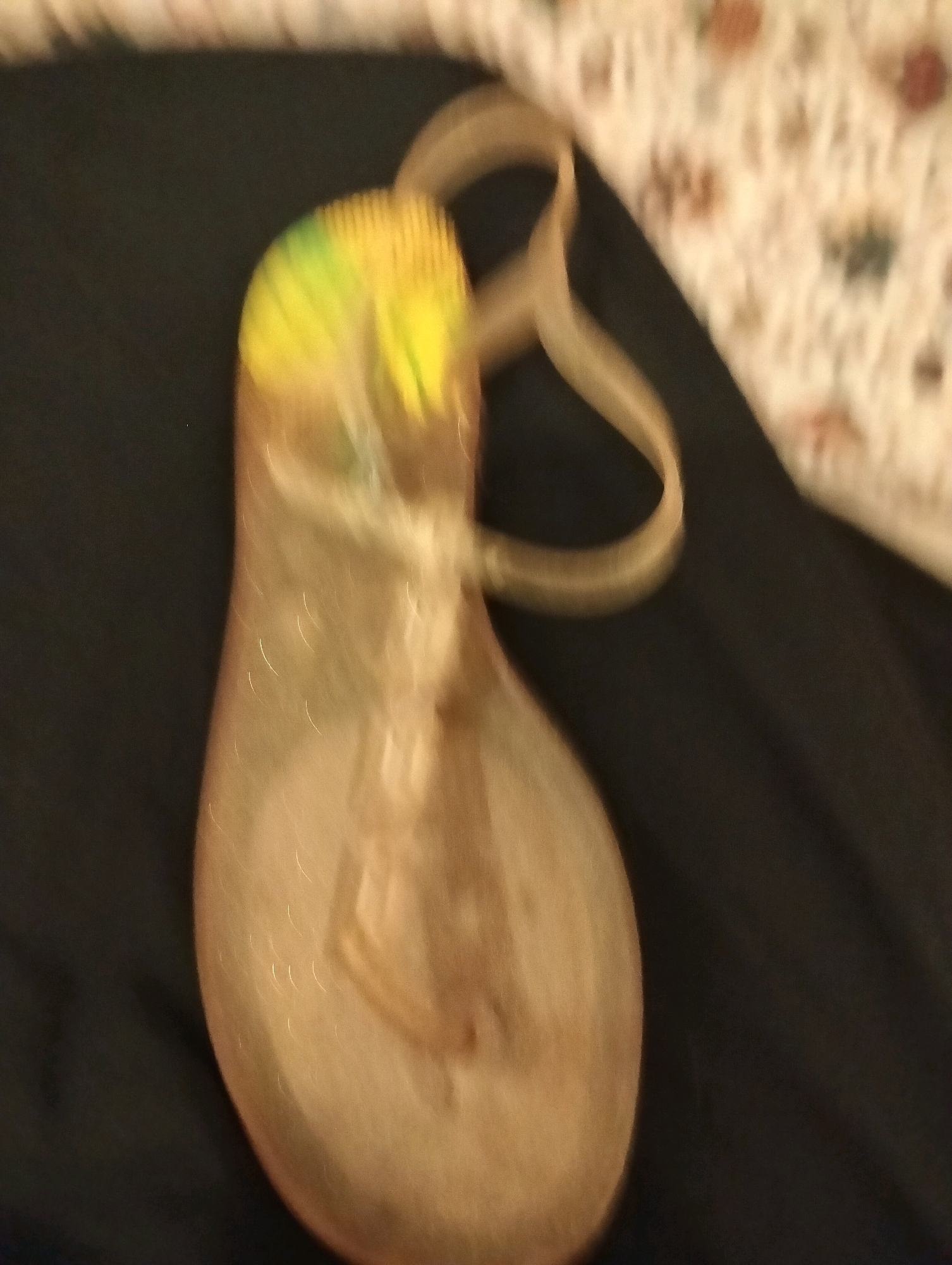 A blurry, top-down view shows a translucent, peach-colored jelly sandal with a yellow and green toe strap, resting on a dark surface. The sandal has a thin strap that loops over the heel. The background is a blurry white surface with a floral pattern and a dark fabric. There are no people or explicit interactions depicted. The scene appears to be indoors. The location in Balçova, Turkey, cannot be determined from the image.