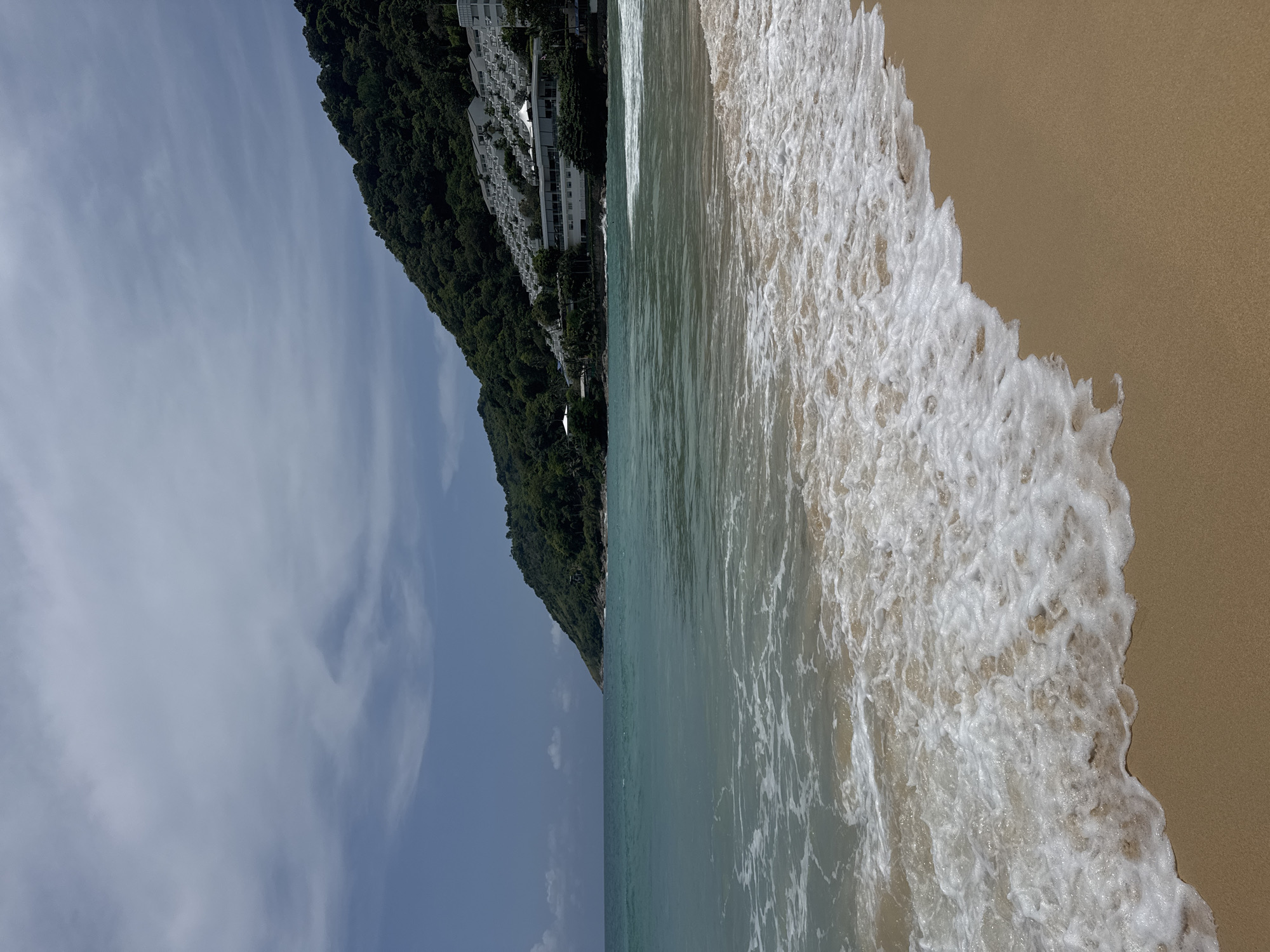 The image shows a beach scene with the ocean in the foreground, a sandy beach, and a green, forested hill in the background. The water is a clear turquoise color with gentle waves lapping at the shore. The beach is made of light brown sand. On the hillside, there are buildings, some of which appear to be hotels or resorts, with white facades and many windows. The sky is partly cloudy, with patches of blue and white clouds. The scene appears to be from Rawai, Thailand, a coastal district known for its beaches and resorts. There are no people visible in the image.