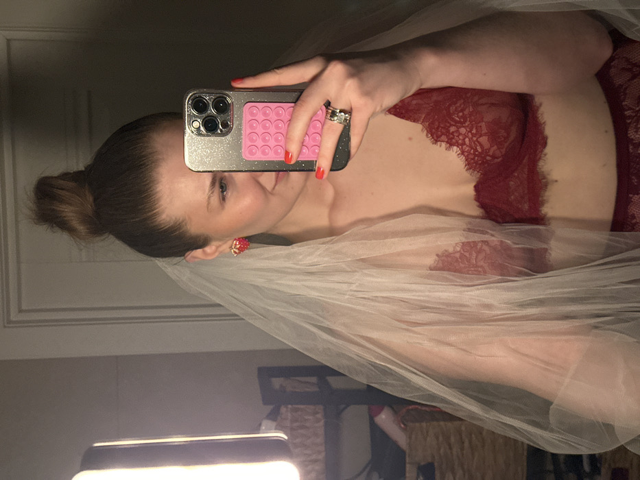 The image displays a female individual taking a mirror selfie indoors, in what appears to be a private residence in Kazan, Russia. The woman, shown from approximately the chest up, has her brown hair secured in a messy bun and wears a sheer white veil draped over her head and shoulders. She is dressed in a deep red lace bralette with matching lower undergarment visible. Red nail polish coats her fingernails. Her left hand holds a silver-colored smartphone, equipped with a glittery case and a pink multi-suction cup grip on its back, partially obscuring her face. Multiple rings are visible on her left ring finger. She wears red, possibly strawberry-shaped, stud earrings. Her visible facial portion shows a slight smile. The background reflection includes a prominent vertical rectangular light source on the left, a white door or wall, a woven basket, and a dark framed object.