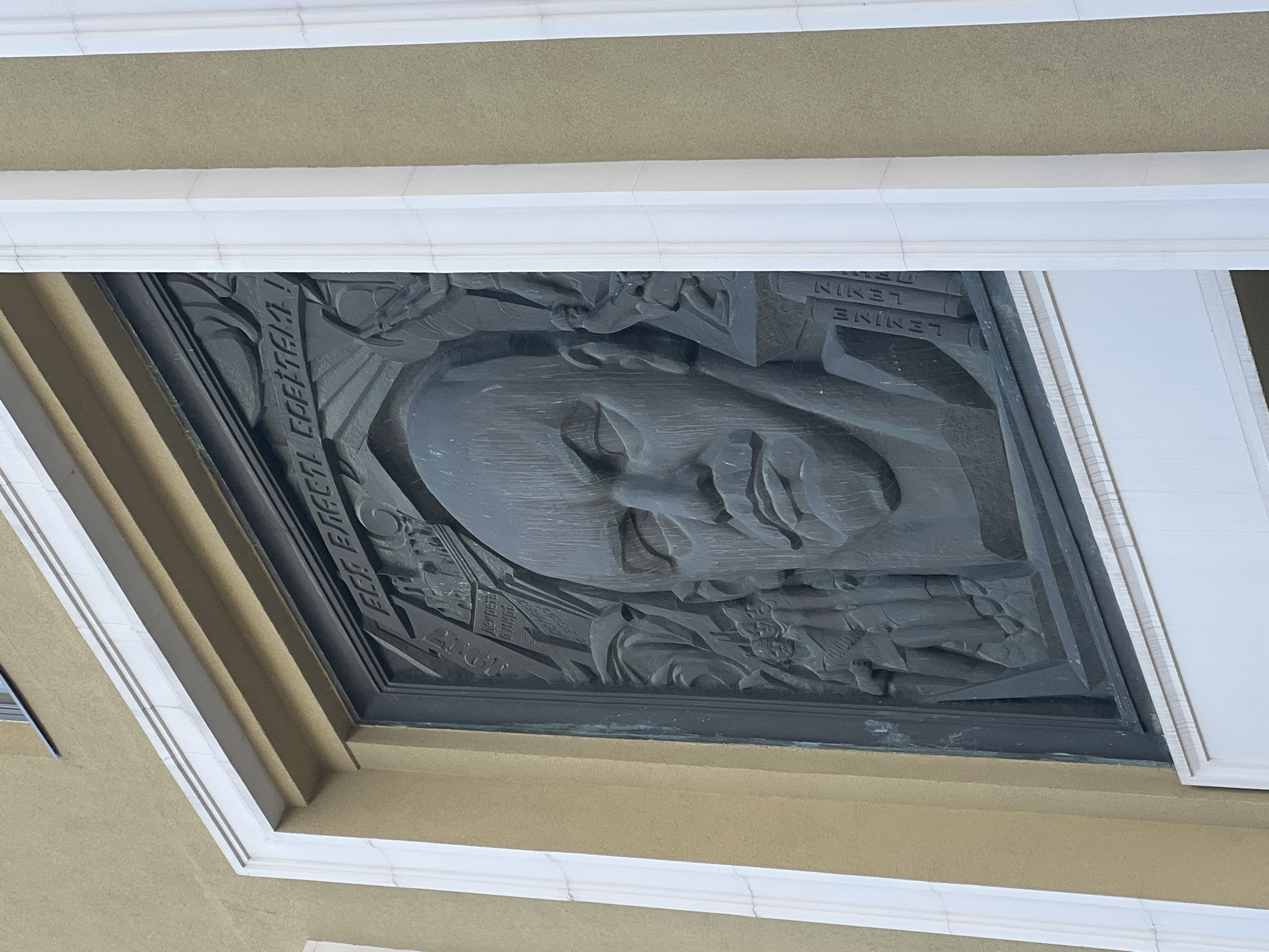 This is a low-angle, close-up shot of a bas-relief sculpture mounted on the facade of a building. The sculpture depicts a portrait of a man in profile, with his gaze directed upwards and to the right. The man's face is rendered in a stylized, somewhat severe manner, with sharp lines and prominent features. He appears to have a stern or determined expression.
Around the central portrait, the relief includes various symbolic elements. To the left of the man's head, there are depictions of books or scrolls, suggesting knowledge or ideology. To the right, there are representations of hands and perhaps gears or machinery, hinting at industry or collective action. The overall composition evokes themes of leadership, progress, and revolution.
The sculpture is made of a dark metal, possibly bronze, and is set within an architectural frame. Above and to the sides of the relief, there are decorative architectural elements, including white moldings and a textured, beige-colored building material. The lighting suggests it is daytime, with soft shadows indicating the sun is not directly overhead.
Visible text in the sculpture is in Cyrillic. On the left side, the inscription reads "ВСЕМ ЕГЕГЕМ!" which translates to "TO ALL THE PEOPLE!". On the right side, stacked vertically, the inscription reads "ЛЕНИН", the Russian transliteration of "LENIN". These inscriptions, along with the portrait and symbolism, strongly indicate that the subject of the relief is Vladimir Lenin, a key figure in the Russian Revolution. The architectural style and the presence of Lenin's name and image suggest a location in Russia, likely Moscow, given the historical significance of Lenin in that city. The scene is one of monumental art intended to commemorate and glorify a historical figure and his associated ideals.