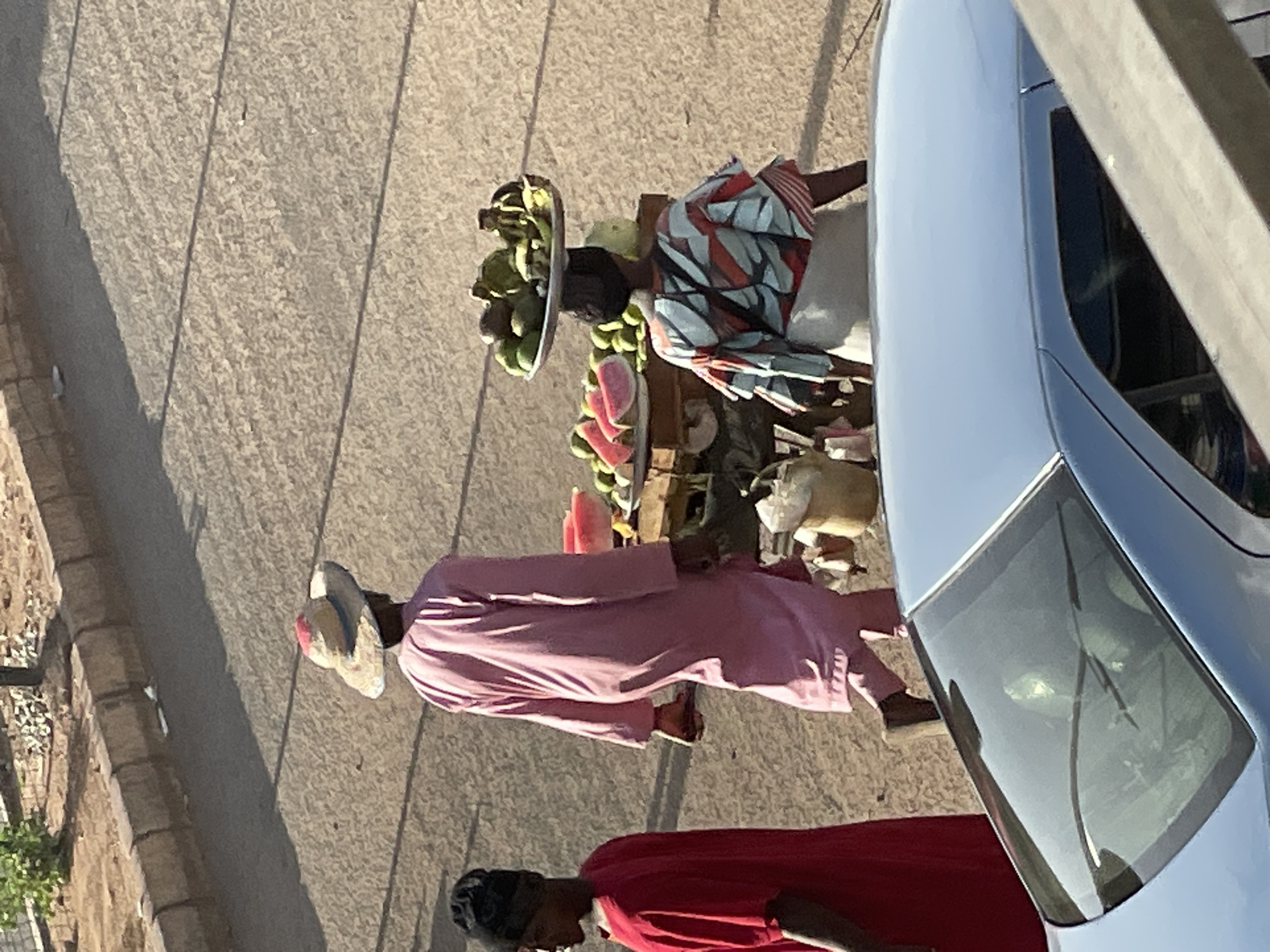 A street in Bukuru, Nigeria. A street vendor stands behind a cart filled with watermelon, bananas, and other produce. A woman with a basket of fruit on her head is walking past the cart. A man in a pink outfit is walking in front of the cart. A parked silver car is to the right of the image.