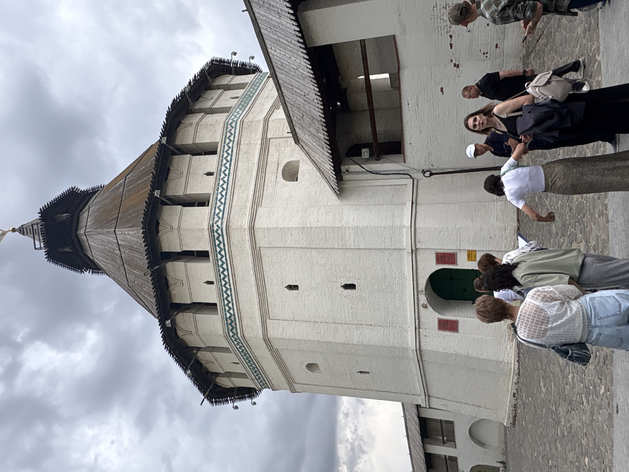 The image depicts the Prechistenskaya Gate Tower of the Astrakhan Kremlin in Astrakhan, Russia, under an overcast sky.
The central subject is a tall, circular, multi-tiered tower constructed of white brick. It is topped with a steep, weathered conical wooden roof, featuring serrated wooden eaves and a small finial at its apex. Below the roof, a series of white, fortified structures with arched openings are visible, above a distinctive green and white geometric patterned band that encircles the tower. The main body of the tower consists of white brick walls, punctuated by several narrow, vertical rectangular openings and a few larger, arched recesses. At the base, a dark green arched wooden door serves as an entrance, flanked by two red rectangular plaques and a smaller yellow sign on the right.
Extending from the right side of the tower, lower white brick walls with dark wooden roof overhangs create open archways and passages, suggesting a fortified perimeter walkway. A similar, less visible wall extends to the left. The ground is paved with irregular dark grey cobblestones.
Approximately ten individuals are present in the lower half of the image. In the mid-foreground, a woman with short brown hair, wearing a white patterned sheer top and light blue jeans, stands with her back to the viewer, facing the green door. To her right, a woman with dark hair, dressed in a light green top and a long grey skirt, also stands with her back to the viewer. Several other individuals are partially visible near this group, some holding papers. To the right, a woman in a long black dress, carrying a black jacket over one arm and a light-colored backpack, is looking directly towards the camera. Adjacent to her, a man in a white baseball cap and black t-shirt faces left. Further right, a man in a plaid shirt and baseball cap is seen from the back, holding a small dark device. Another man in a black t-shirt is partially visible behind the woman in black. The individuals appear to be gathered near the tower's entrance, possibly as part of a tour or visit.