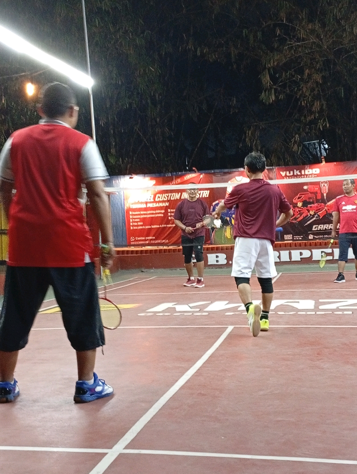The image shows a badminton court in Caringin, Indonesia. Four men are playing badminton under artificial lights. The court surface is red, with white lines. The men are wearing athletic clothing. One man in the foreground is wearing a red and white shirt and dark shorts, holding a badminton racket. Another man in the middle ground is wearing a maroon shirt and white shorts, also holding a racket. To the left, a man in a maroon shirt and black shorts is visible, and to the right, a man in a red shirt and dark shorts is also present. In the background, a large banner with text in Indonesian and English, including "YUKIDO" and "CUSTOM INDUSTRI," is displayed. Bamboo plants are visible behind the banner. The lighting suggests it is nighttime.