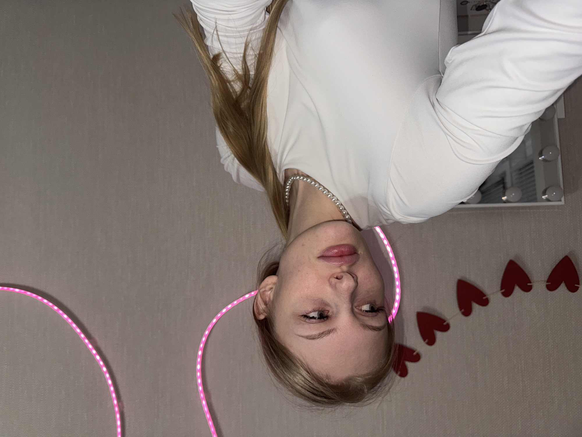 This indoor portrait features a young woman with long blonde hair, fair skin, and light-colored eyes, captured from the chest up. She wears a white long-sleeved top and a delicate pearl necklace. Her head is slightly tilted, and her gaze is directed towards her right, away from the camera, with a calm and contemplative expression and slightly parted lips. Her hair falls over her shoulders.
Bright pink LED strip lights curve on both sides of her, partially framing her face and head. In the background, to her right, a string of red heart-shaped decorations forms a garland across a light gray, textured wall. A portion of a vanity mirror with illuminated circular bulbs is visible behind her, towards the upper right corner of the frame. The artificial lighting, primarily from the pink LEDs and the mirror, suggests an intimate, evening setting. The overall atmosphere is personal and relaxed, likely in a private room in Perm, Russia. No visible text is present in the image.