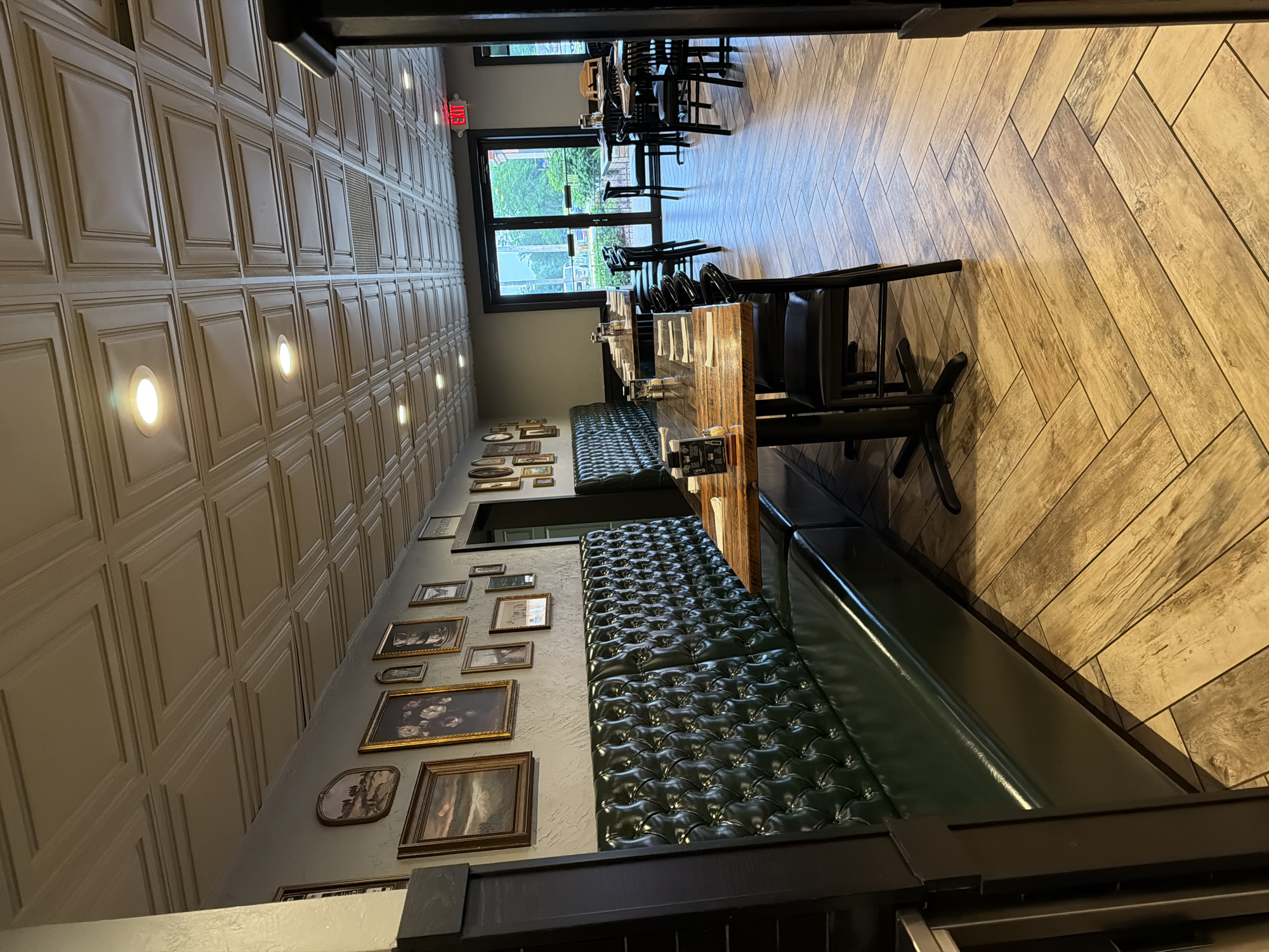The image shows the interior of a restaurant in Lake Delton, Wisconsin, United States of America. The restaurant features dark green tufted booth seating along the left wall, with several wooden tables and chairs set up throughout the room. The tables are adorned with white napkins and menus. The floor is laid with light brown wooden planks arranged in a herringbone pattern. The ceiling is paneled with square coffers, and recessed lighting fixtures are visible. Framed artwork hangs on the grey walls. In the background, a glass-paneled door leads to an outdoor area with greenery visible beyond. An "EXIT" sign is illuminated above the door.