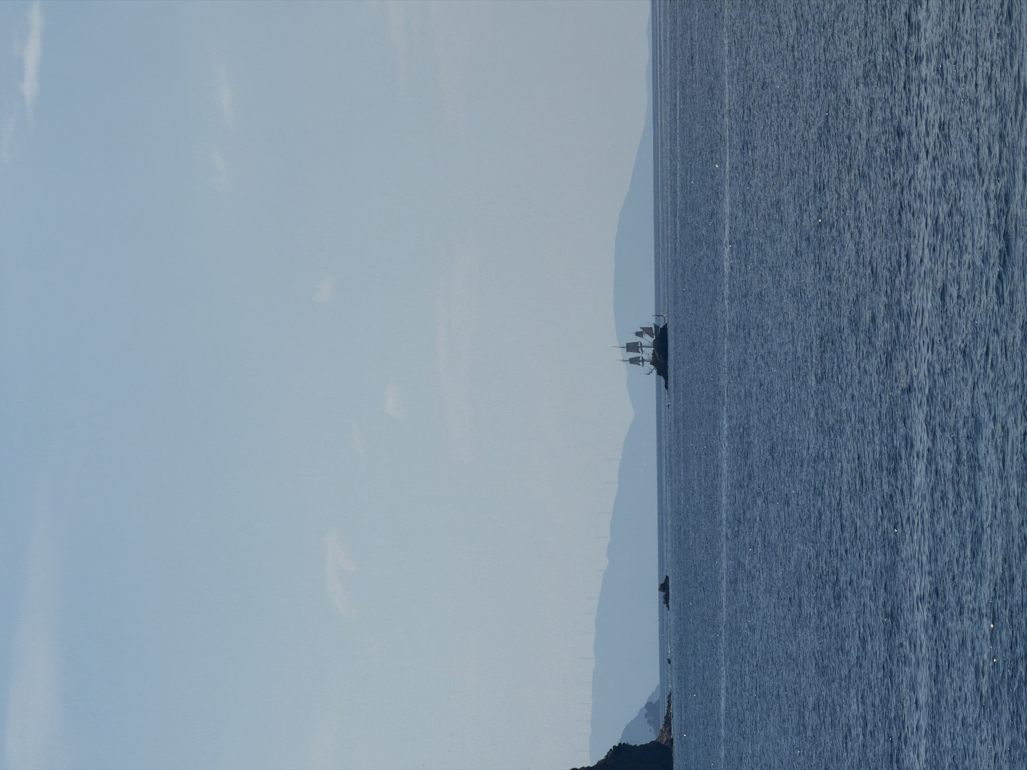 The image captures a scenic view of the Adriatic Sea, likely taken near Budva, Montenegro. The foreground consists of a vast expanse of ocean with gentle ripples. A dark land mass borders the ocean. In the distance, a faint, undulating mountain range is partially obscured by a hazy sky. A sailing vessel, resembling a traditional sailing ship, can be seen near the center of the frame, just offshore. The sky above is a pale, almost milky, blue, suggesting a bright but possibly overcast day. The overall color palette is dominated by blues and grays, creating a calm and serene atmosphere.