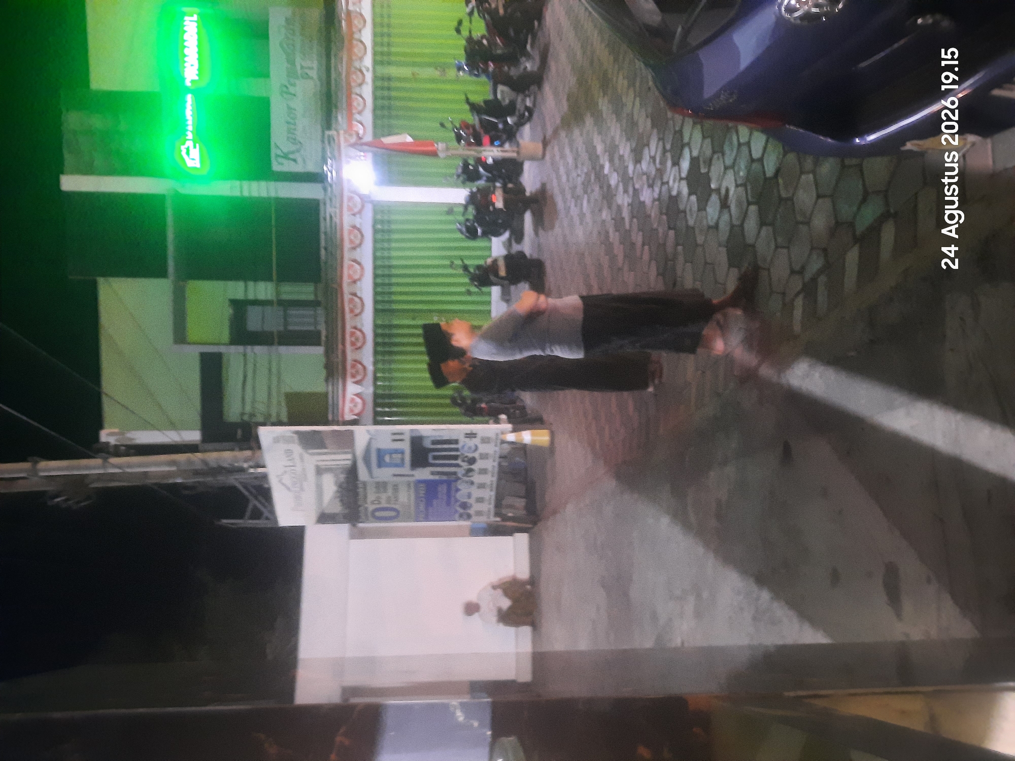 In Sukokerto, Indonesia, at 19:15 on August 24, 2026, an exterior night scene depicts a building with a bright green neon sign. The sign displays text in what appears to be Indonesian, including "PRADAMAIN." Below the neon sign, a white sign reads "Kantor Pemasaran PT." In front of the building, a group of men wearing traditional Islamic attire, including peci (caps), are standing or observing. One man in the foreground is wearing a grey t-shirt and dark pants, with his arms crossed. Behind them, several motorcycles are parked. To the left of the scene, a man in a white shirt and brown pants is seated on the ground, leaning against a white wall. The ground is paved with hexagonal stones. A blue car is visible in the lower right corner of the image.