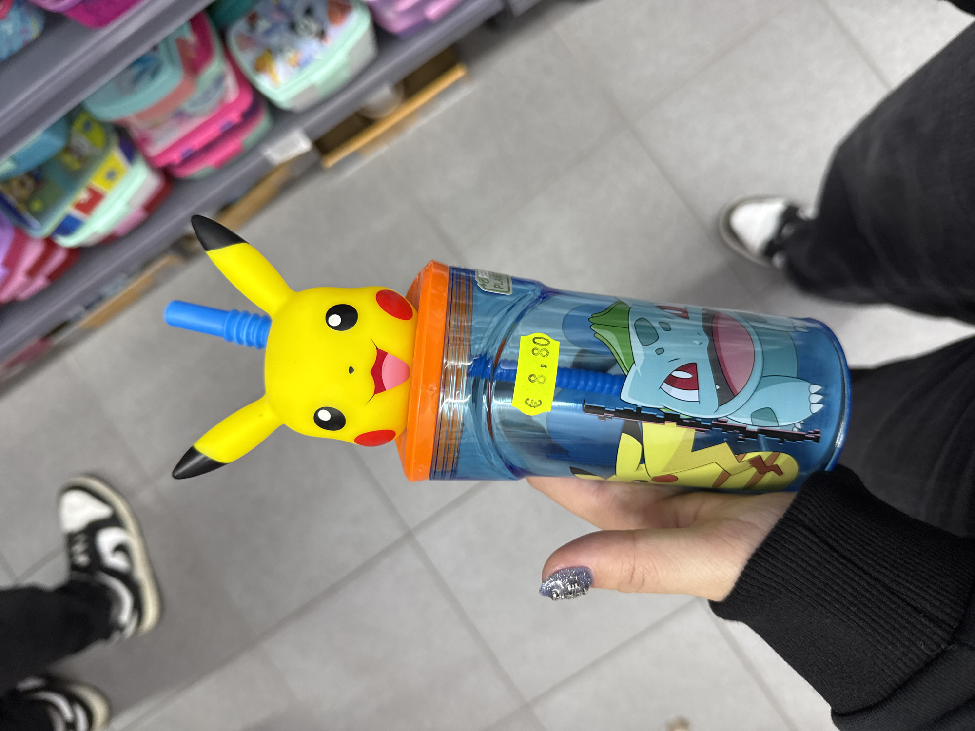The image shows a person holding a blue Pokemon-themed cup with a yellow Pikachu head on top. The cup has images of other Pokemon like Bulbasaur. A yellow price tag of €8.80 is on the cup. The person's hand holding the cup has a glittery nail design. The background includes shelves of lunchboxes in various colors and designs. The person is wearing black pants and white shoes with black details. The scene is indoors, likely in a store. There is no evidence to indicate the location is Madrid, Spain.