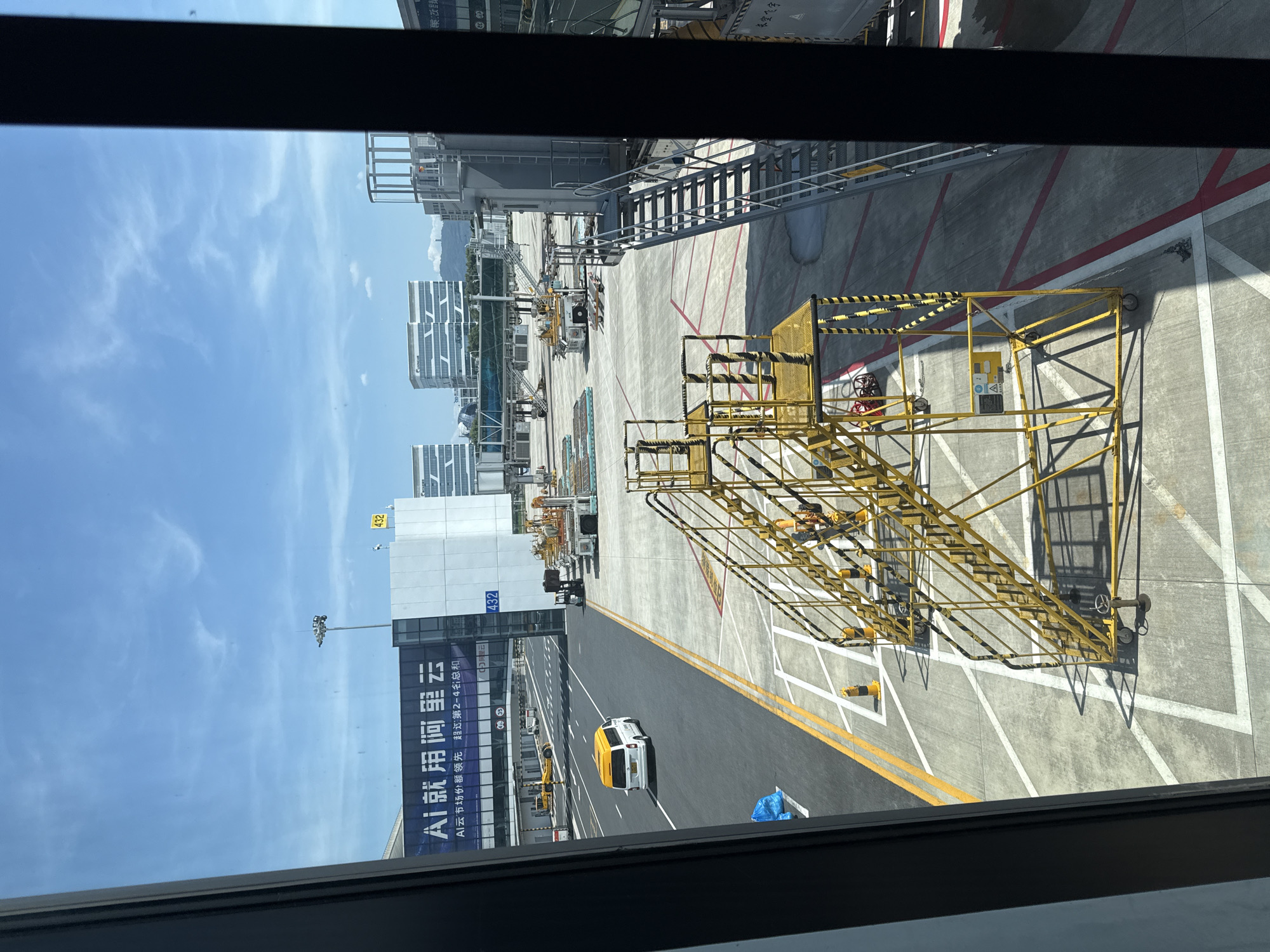 This image captures a view of an airport tarmac, likely from inside an airport terminal or a vehicle. In the foreground, a bright yellow, multi-tiered mobile staircase with black and yellow striped safety railings is prominently positioned. It has wheels for mobility and a steering wheel at its base. To the left of the tarmac, a white van with a yellow roof is driving on a road. A blue plastic bag lies on the side of the road near the van. In the middle ground, various airport vehicles and equipment are visible, including service carts and a small yellow tug. A pedestrian bridge, possibly connecting terminals, stretches across the tarmac. Further in the background, modern, multi-story buildings with distinctive architectural designs stand against a clear blue sky dotted with wispy clouds. One building prominently displays a large banner with Chinese text that reads "AI 就用阿里云" (AI, use Alibaba Cloud) and other smaller Chinese characters. A yellow sign with the number "432" is attached to a building structure. The environment appears to be an active airport operational area. The scene is illuminated by bright daylight, casting shadows on the tarmac. The location is identified as Xiaoshan District, China, suggesting this is a view from Hangzhou Xiaoshan International Airport.