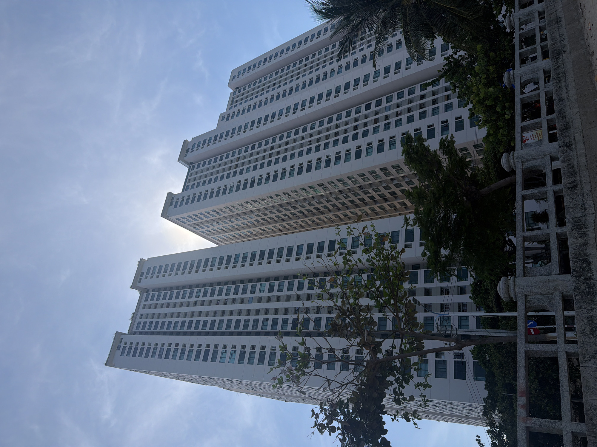 The image captures a low-angle perspective of two modern, white high-rise buildings located in Bac Nha Trang Ward, Vietnam. The buildings exhibit a stepped design with multiple floors of windows. Sunlight is visible behind the buildings, creating a bright glow in the sky. Some greenery, including trees and palm leaves, partially obscures the view of the buildings on the right side. A gray concrete fence is in the foreground. The sky is pale blue with some scattered clouds. The image has a tilted orientation, suggesting it was taken while the camera was angled upwards.