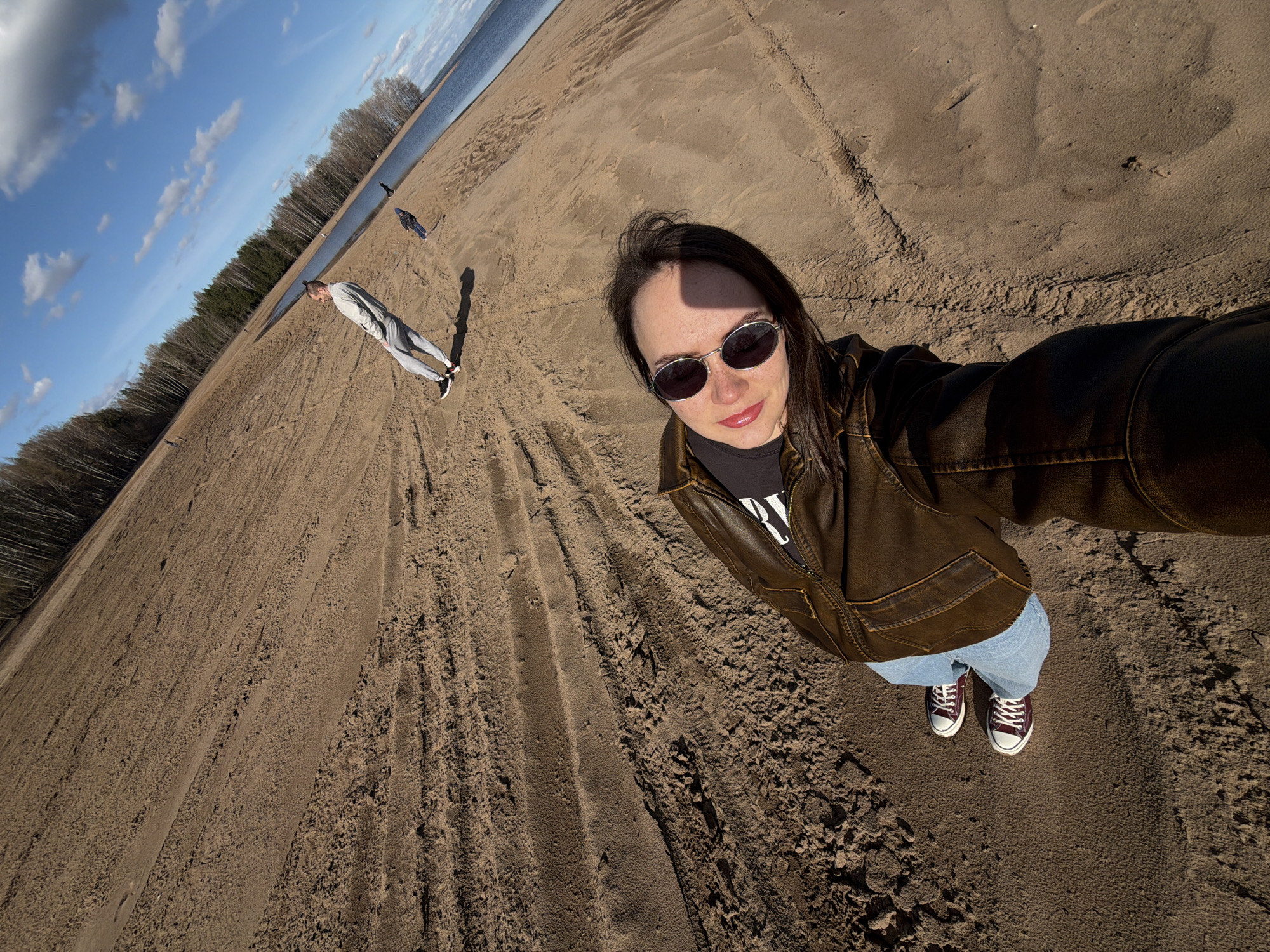 The image is a selfie taken at a beach in Chaikovsky, Russia. The photographer, a woman with dark hair and sunglasses, is wearing a brown leather jacket, jeans, and maroon sneakers. In the background, a man in a grey tracksuit is walking on the beach, and there are other people further away. The sky is blue with some clouds, and a forest line is visible in the distance. The beach is sandy and appears to be alongside a body of water.