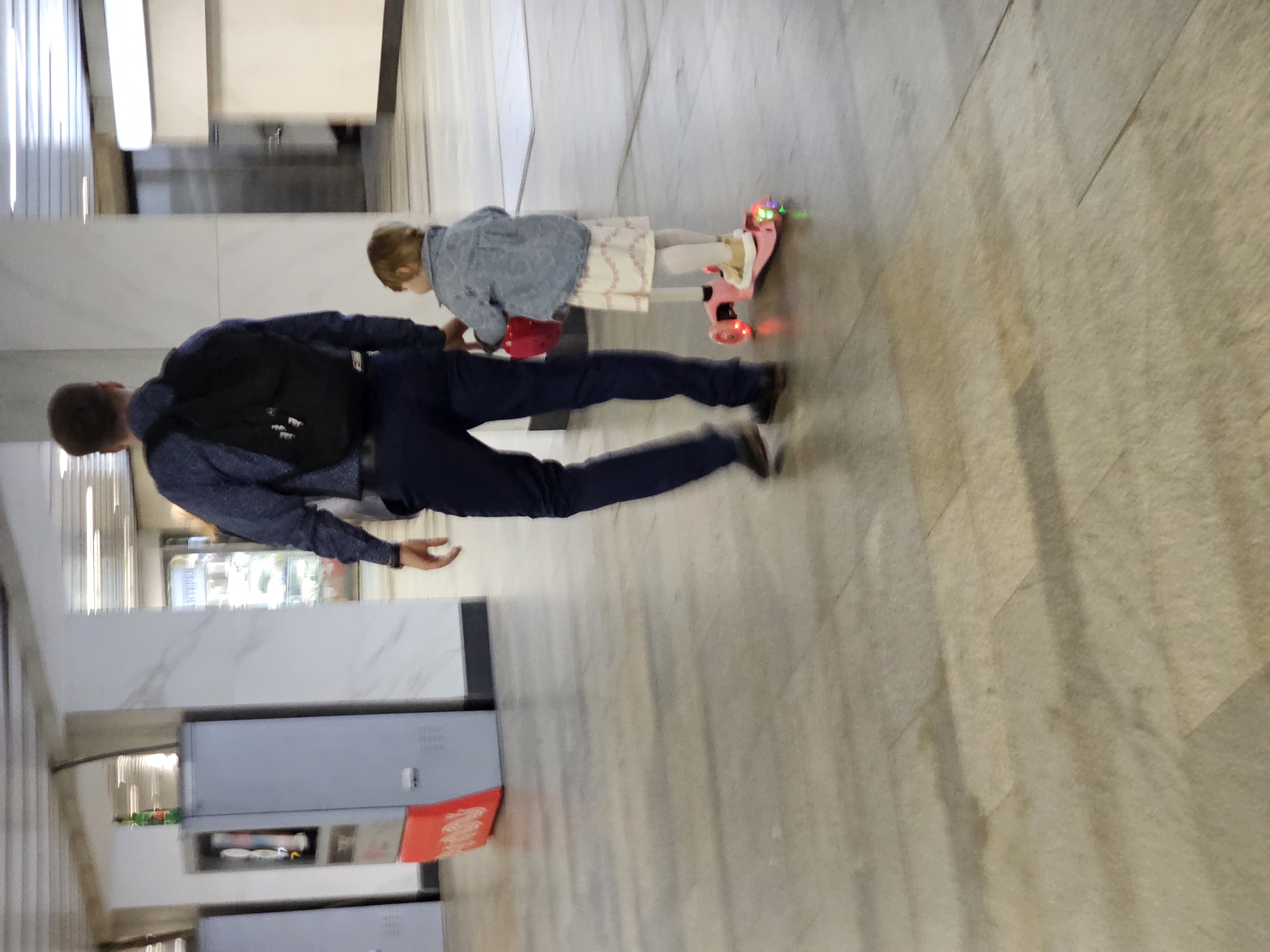 A man in a dark blue button-down shirt and dark pants walks ahead of a young girl on a pink scooter. He has a black backpack on. The girl is wearing a light blue denim jacket over a white dress with pink trim and white tights. She is holding a red object in her left hand. The scooter has flashing colored lights on its wheels. They are in a well-lit indoor space with a tiled floor. In the background, there are vending machines and a large pillar. The overall environment suggests a public indoor space like a subway station or a shopping mall.