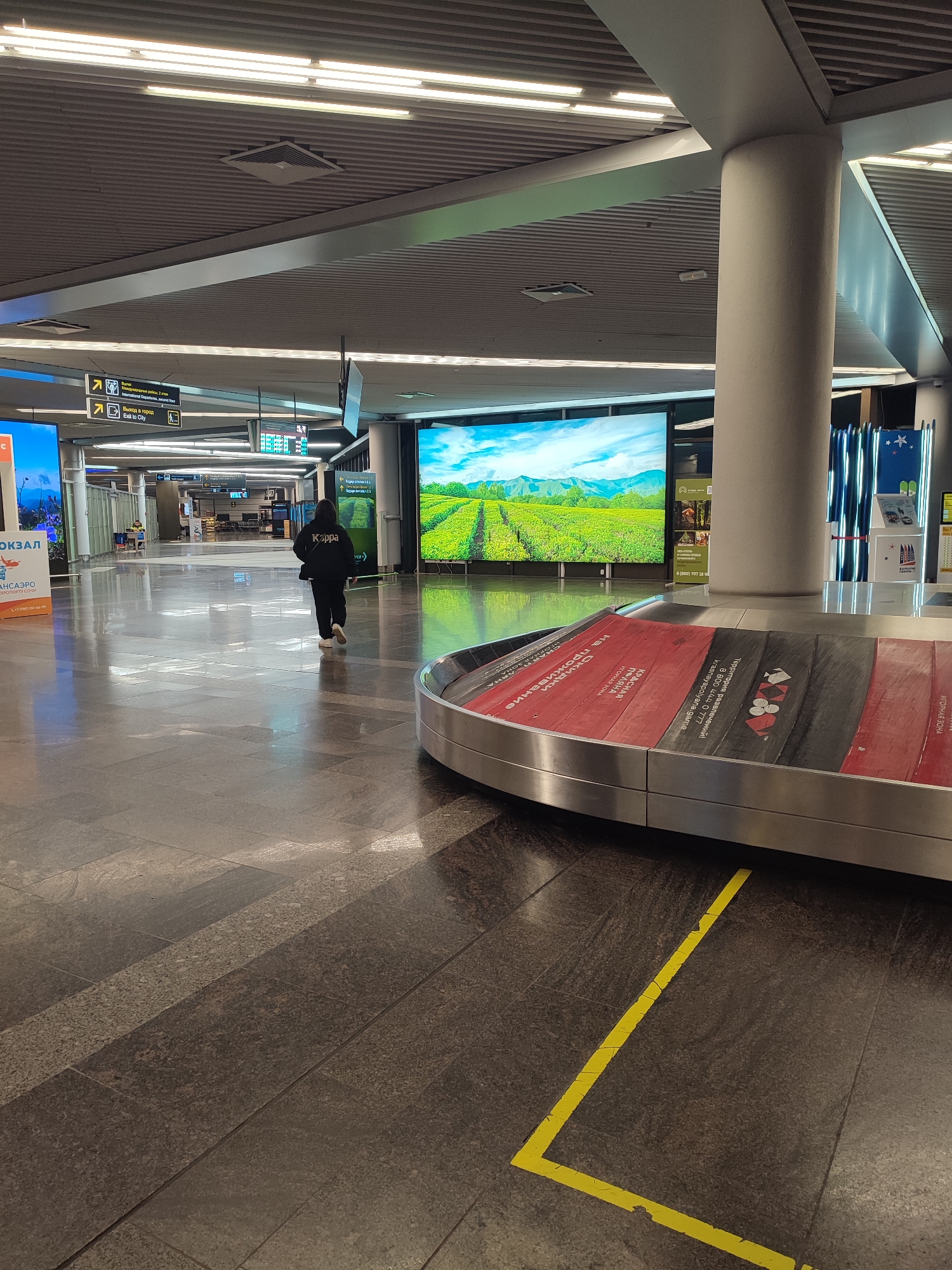The image displays the interior of the Sochi International Airport (AER) in Sochi, Russia, specifically within an arrivals or baggage claim area. The scene is brightly illuminated by numerous overhead fluorescent strip lights integrated into a corrugated metal ceiling. The floor is composed of large, polished dark grey and brown tiled granite, with a yellow L-shaped tape line marked on the floor in the foreground right.
In the mid-ground, a semicircular baggage carousel with a silver-colored perimeter is visible. Its conveyor belt segments are alternately red and dark grey, bearing white Russian text that includes "КРАСНАЯ ЗОНА" (RED ZONE) and warnings such as "НЕ ПРОХОДИ" (DO NOT ENTER). To the left of the carousel, a single individual is walking away from the viewer, moving towards the left side of the frame. This person is dressed in a black hooded sweatshirt with the word "Kappa" emblazoned in white on the back, black pants, and white shoes, carrying a dark bag over their left shoulder.
The background features a large, rectangular LED screen displaying a vibrant image of green, terraced fields or tea plantations under a blue sky with white clouds and distant mountains. To the left of this screen, a large, light-colored cylindrical pillar extends from the floor to the ceiling. Further to the left, yellow and black directional signs in both Russian and English are mounted overhead, with one indicating "Выход в город / Exit to City." A partially visible digital display on the far left shows Russian text, including "ВОКЗАЛ" (TRAIN STATION). The area appears clean and sparsely populated.