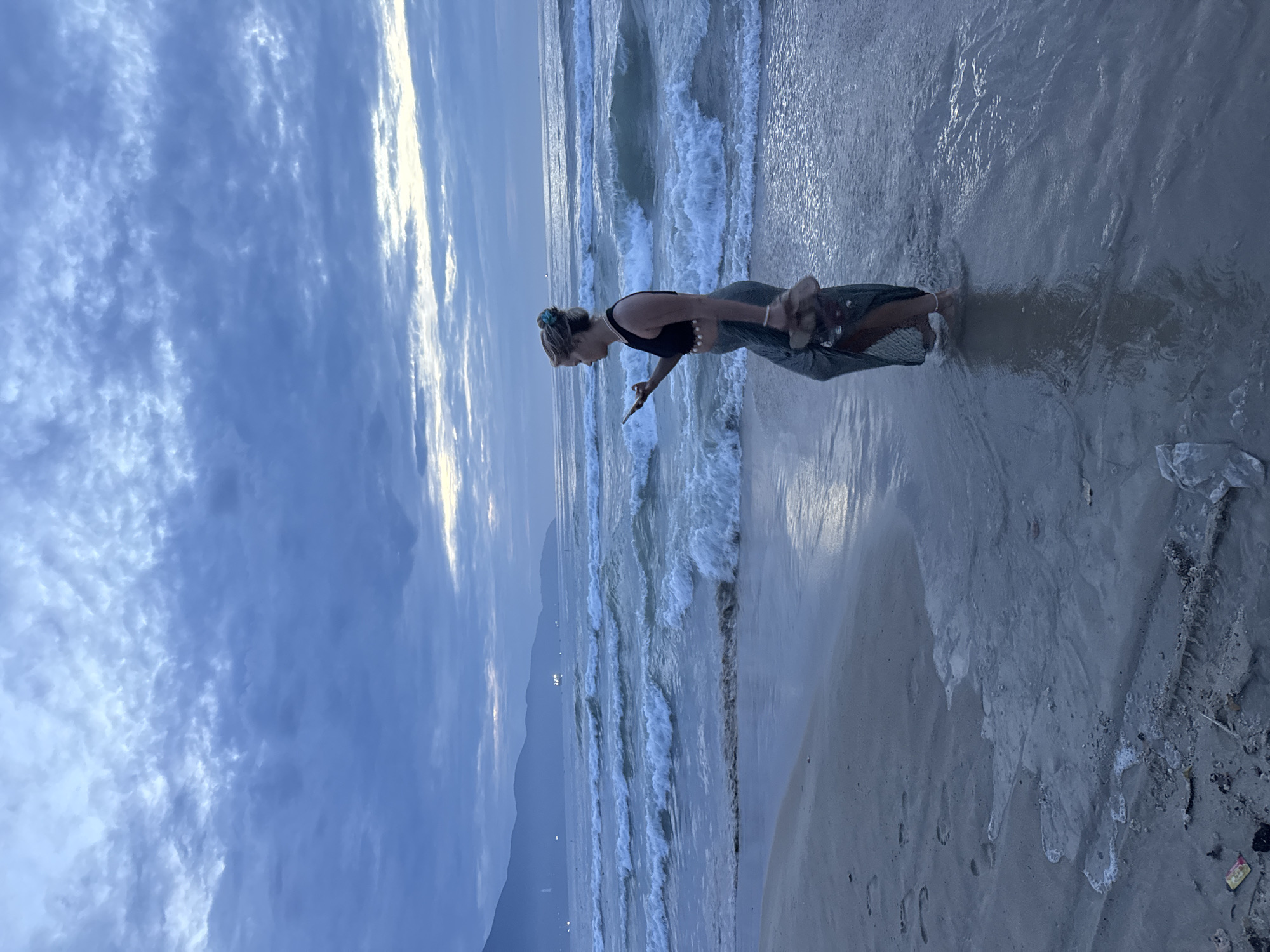 The image depicts a woman standing on a beach at dusk or dawn. The woman is holding a mobile phone in her hand, looking at the screen. She is wearing a black top, a long gray skirt, and sandals.
The location is the city of Đà Nẵng, Vietnam. The scene shows a beach, waves coming in and out, a cloudy sky, and some distant hills/mountains in the background. The light suggests that it is either sunrise or sunset. There is also some litter visible on the sand.