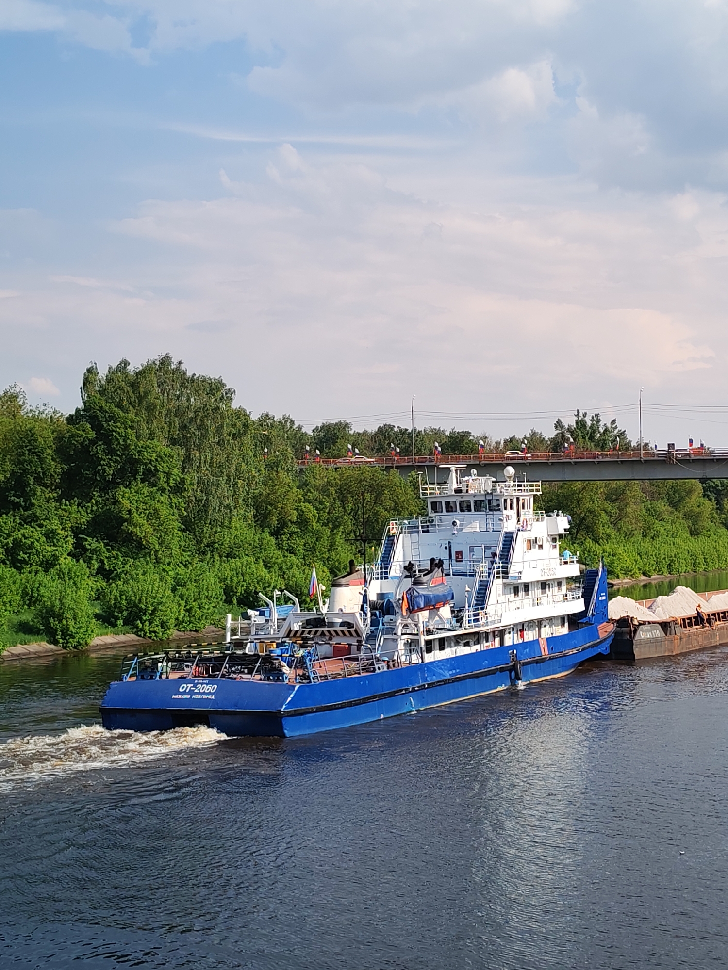 The image captures a blue and white river tugboat, the "OT-2060", navigating a river in Yakhroma, Russia. The tugboat is towing a barge filled with a light-colored cargo, possibly sand or gravel. Lush green trees line the riverbank, and a bridge spans the river in the background, adorned with small flags along its length. The water is dark and reflective, showing the wake of the tugboat. The sky above is partly cloudy, with patches of blue visible.