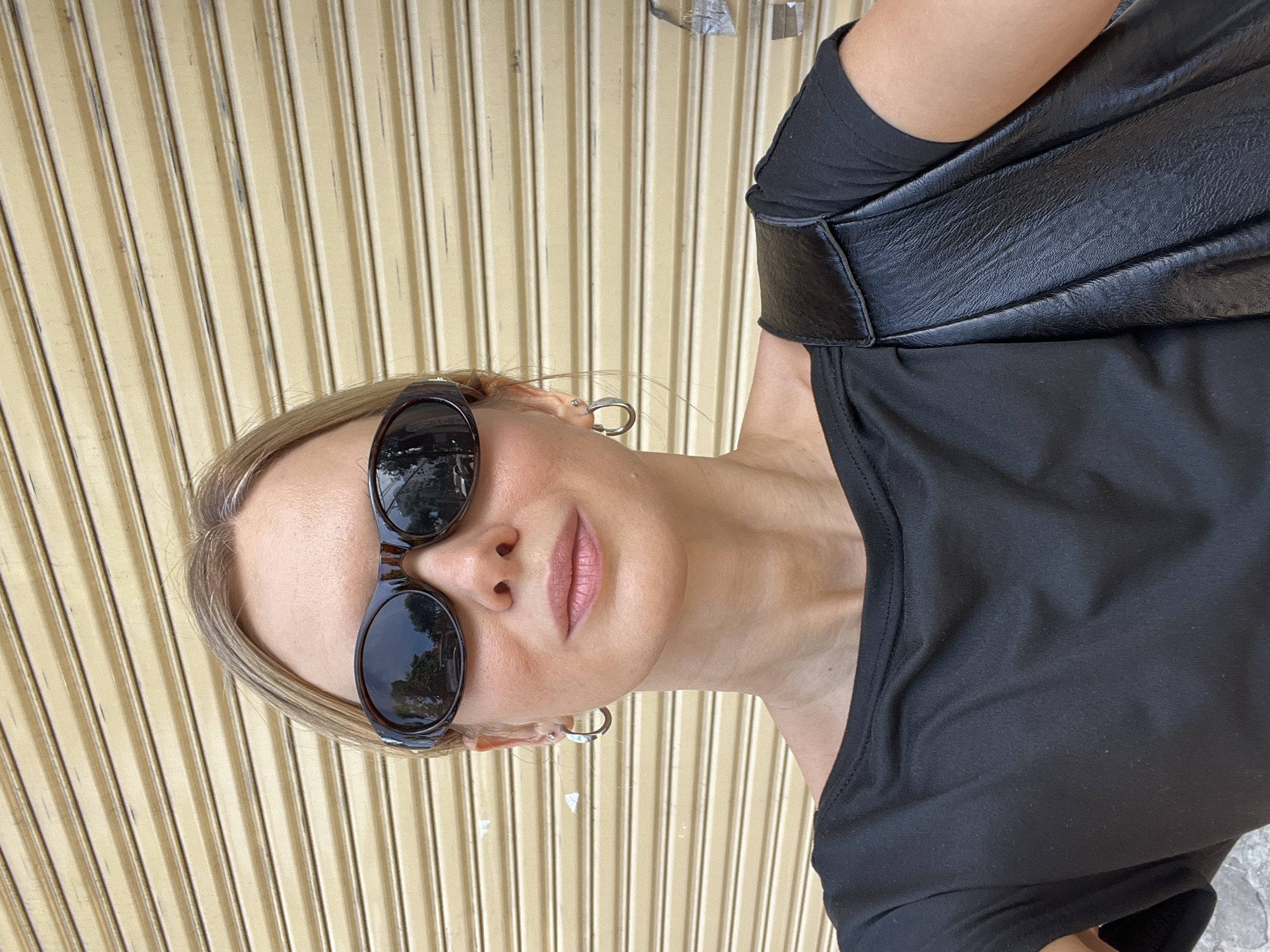 A woman with blonde hair pulled back is standing outside, facing the camera. She is wearing dark sunglasses, a black t-shirt, and silver hoop earrings. A black leather bag is slung over her right shoulder. Behind her is a ribbed, beige metal door, likely a shop shutter. The setting is outdoors on a street in Nha Trang, Vietnam.