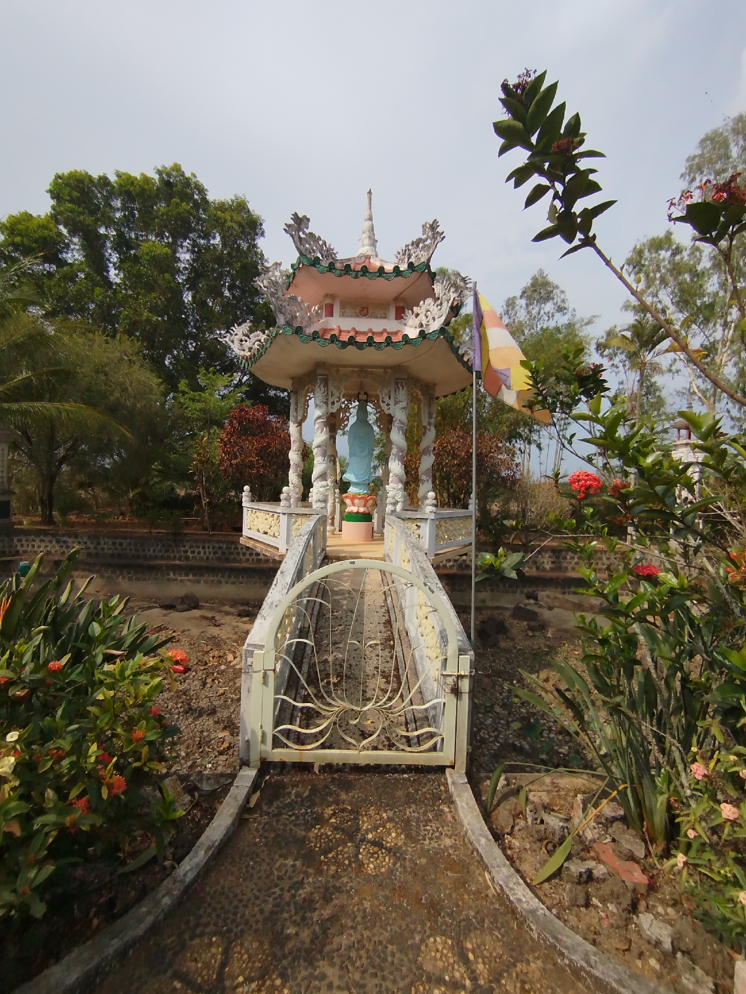 The image shows a small, ornate pavilion in Phú An, Vietnam. The pavilion, with a multi-tiered roof, is situated on a raised platform and houses a blue statue. A small bridge with a gate leads to the pavilion. The surrounding area appears to be a garden with various plants and trees. A Buddhist flag flies near the pavilion. The foreground is filled with vegetation, including flowering bushes. The sky is overcast.