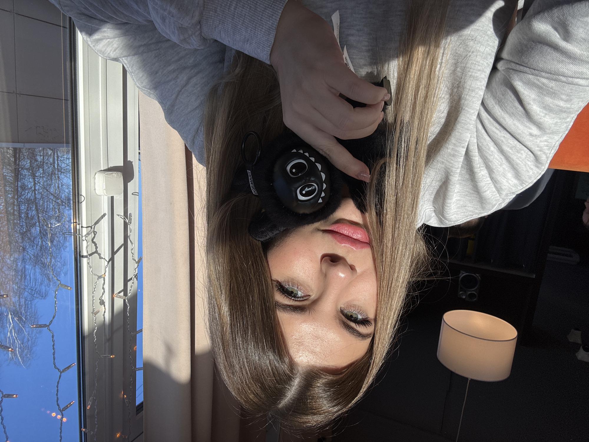 A young adult female, facing forward, occupies the foreground of an indoor scene. She has light brown hair, green eyes, and pink lipstick, wearing a grey crewneck sweatshirt. Her left hand holds a black plush object, possibly an earmuff or small toy, featuring prominent white eyes and teeth, with "POP MART" branding visible. Strong direct sunlight from the right illuminates her, casting shadows. In the background to the left, a dark ceiling with a white drum-shaped pendant lamp and a black speaker are visible, along with an orange upholstered surface. To the right, a large window reveals a clear blue sky and bare deciduous trees outside. Vertical strands of warm-toned string lights are affixed to the window pane. A beige curtain is partially visible on the left side of the window frame. No specific visual identifiers are present to precisely locate the scene within the city of Chaikovsky, Russia.