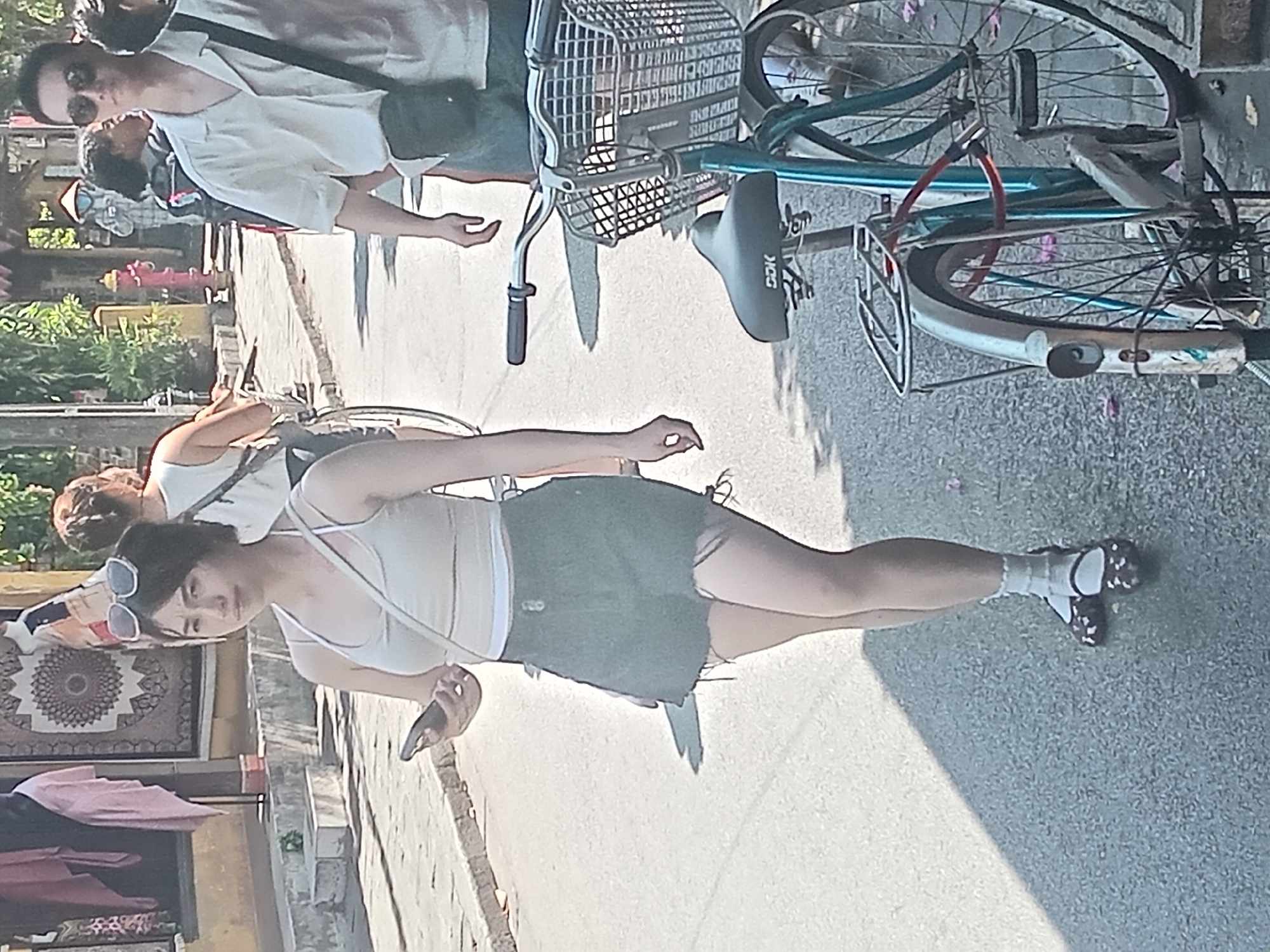 The image depicts a street scene in Hoi An, Vietnam, on a sunny day. In the foreground, a young woman walks towards the viewer, holding a smartphone in her left hand. She is wearing a light-colored tank top, a dark denim skirt with a frayed hem, white ankle socks, and patterned flat shoes. Sunglasses are perched on top of her head. To her right, a light blue bicycle with a front basket is parked, and a portion of a second bicycle is visible behind it. In the background, several people are walking, and a man in a light-colored short-sleeved shirt and sunglasses is walking away from the viewer. The street is paved, and buildings with traditional architecture line the left side of the street. Some foliage and trees are also visible.