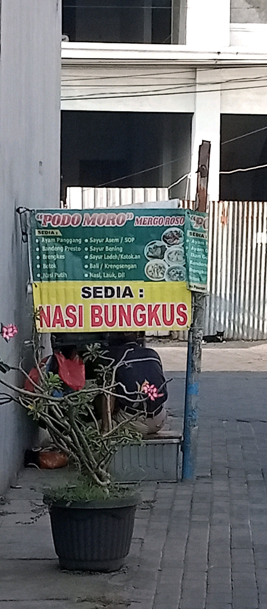 A horizontal, medium-shot photograph captures a street-level view of a food stall in Kepuh Kiriman, Indonesia. The stall features a prominent sign with a dark green background at the top, displaying "PODO MORO" in large white letters, followed by "MERGO ROSO" in smaller white letters. Below this, a yellow banner announces "SEDIA:" in black text, followed by "NASI BUNGKUS" in large red letters. The green sign lists various food items such as "Ayam Panggang," "Bandeng Presto," "Brengkes," "Betok," "Nasi Putih," "Sayur Asem / SOP," "Sayur Bening," "Sayur Lodeh/Kotokan," "Bali / Krengsengan," and "Nasi, Lauk, Dll." Several small circular images of food are printed on the right side of the green sign.
In the foreground, a large dark pot contains a leafy green plant with pink flowers. To the left of the plant, a person wearing a dark blue jacket with white trim and tan pants is seated. Red and orange plastic bags are visible behind the seated person, near the base of the plant. A dark-colored trash receptacle is also present. The background shows a building with dark windows and a light-colored facade, as well as a metal fence with vertical slats. The ground is paved with interlocking gray bricks. A black and white cat is visible walking on the paved area in the distance to the right.