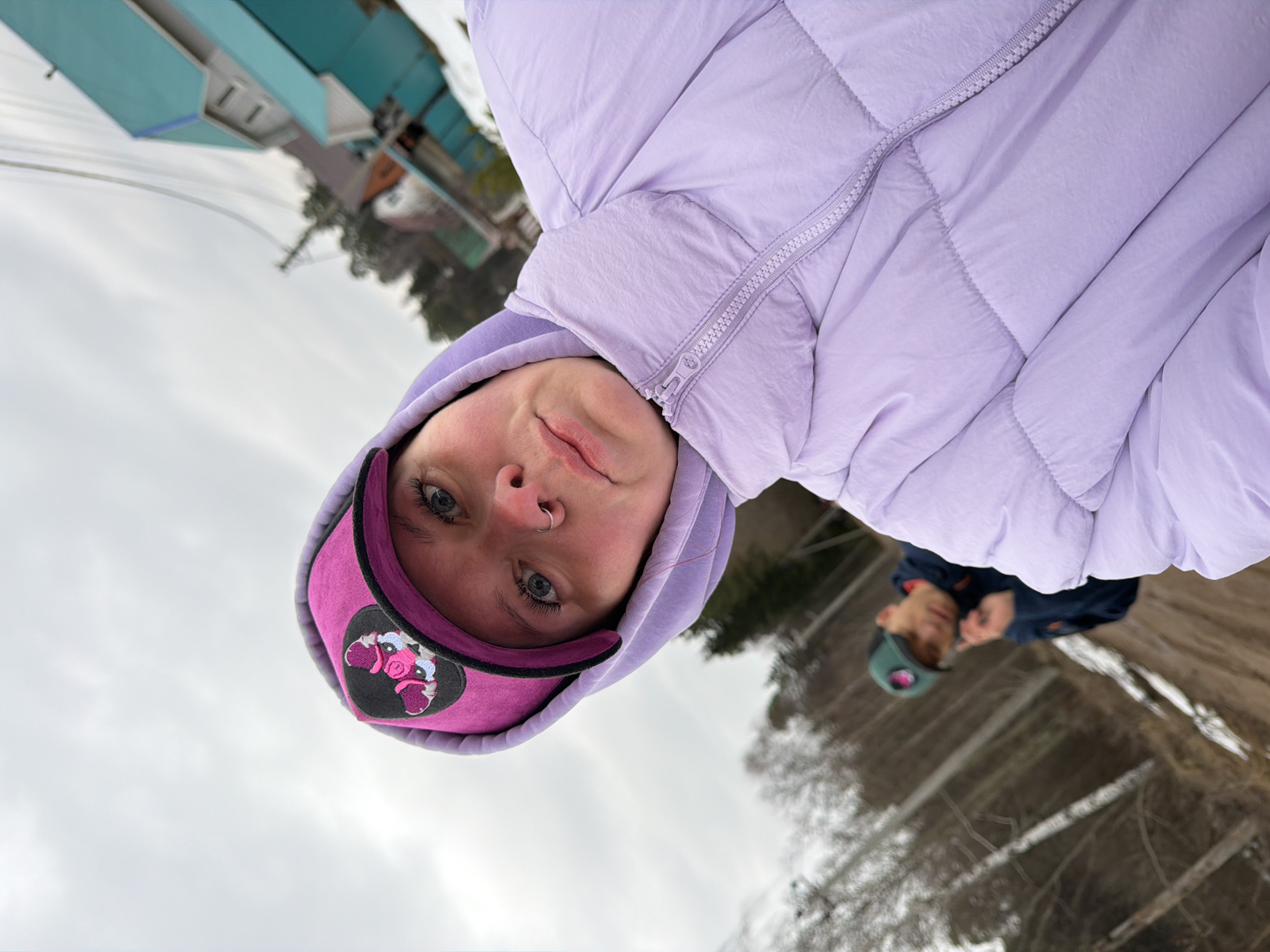 A young person wearing a lilac puffer jacket and a pink baseball cap is pictured. The image appears to be a selfie taken outdoors on a cloudy day. In the background, another person wearing a navy jacket and a green baseball cap can be seen. Trees and snow patches are also visible. The location is in Chaikovsky, Russia.