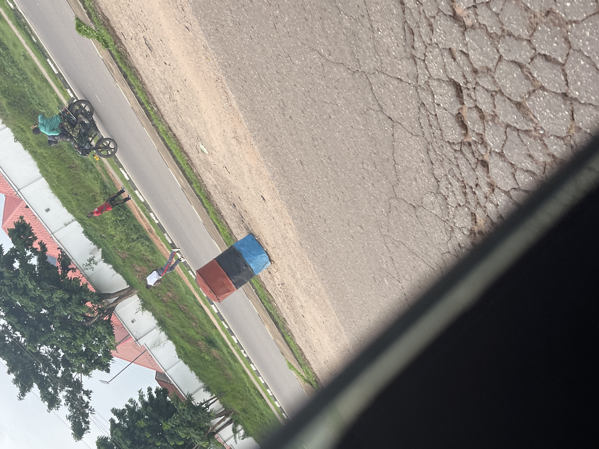 In Aso, Nigeria, a motorcyclist wearing a green jacket and camouflage trousers rides a black motorcycle with a passenger in similar attire. They are traveling on a paved road beside a grassy embankment. To their left, a person in a red shirt stands on the side of the road. Further left, a person in a white shirt and grey pants walks along the road. A cylindrical object painted red, black, and blue stands upright on the side of the road. In the background, trees and buildings with red roofs are visible. The sky is overcast.