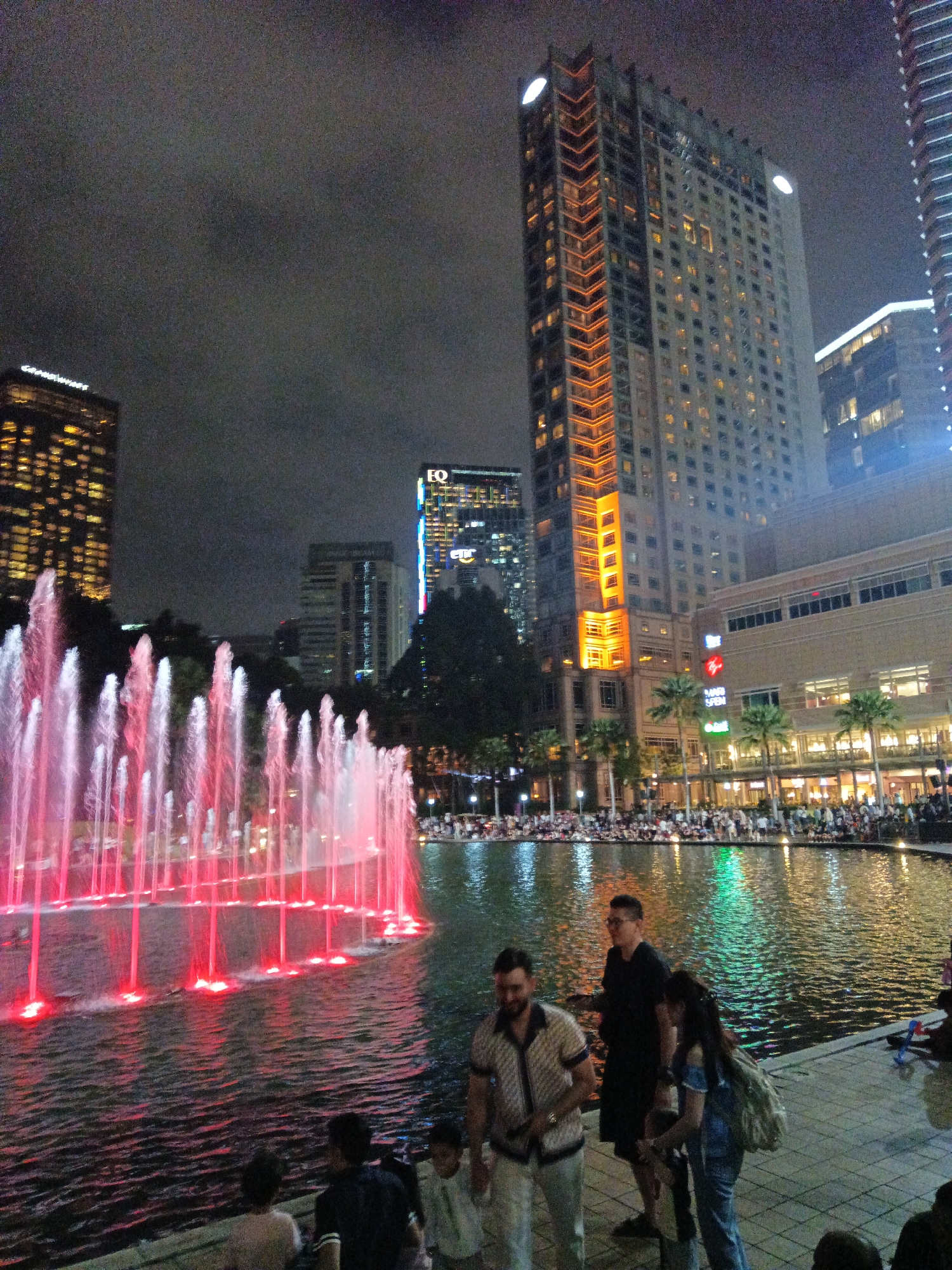 The image depicts the Suria KLCC Twin Towers and its surrounding area at night. A large, illuminated fountain is in the foreground, with water jets lit in red and pink. Several people are visible in the foreground, some standing and watching the fountain, others walking along the edge of the water. In the background, the iconic Petronas Twin Towers and other skyscrapers are lit up, along with a large crowd of people gathered around the fountain and the water's edge. The sky is dark and cloudy. The scene suggests a public gathering or event at a popular tourist destination.