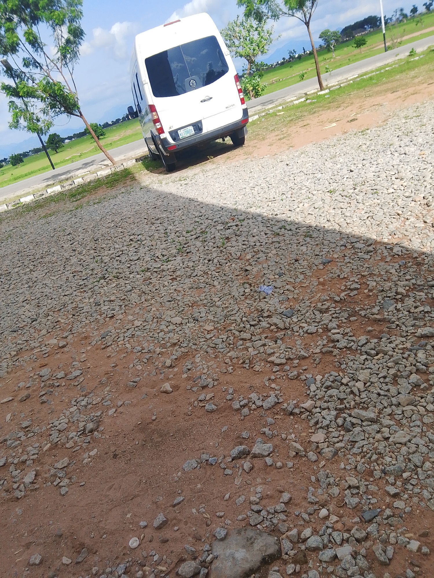 A white minibus is parked on a gravel surface in Jalingo, Nigeria. The minibus is positioned at an angle, with its rear facing the viewer and slightly to the left. The vehicle has tinted windows and a license plate that reads "XU4-786DX". The ground is covered in a mix of small gray stones and reddish-brown dirt. In the background, there is a green grassy field with scattered trees and a road. The sky is blue with some white clouds, suggesting it is daytime. A shadow of a tree falls across the gravel surface in the foreground. The scene appears to be an outdoor setting, possibly a parking area or a roadside. There are no people visible in the image. The overall impression is one of a bright, sunny day in a semi-rural or suburban environment.