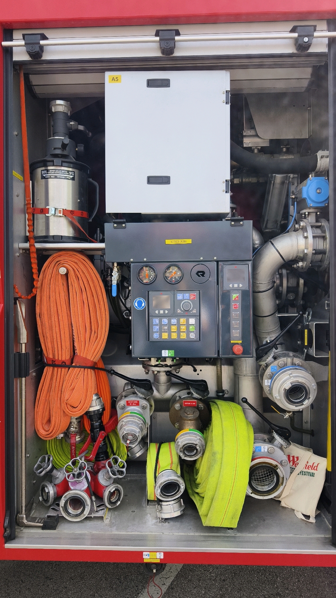The image shows a detailed view of the equipment compartment of a red fire truck, likely located in Vösendorf, Austria. The compartment is filled with various fire-fighting tools and apparatus. Prominently displayed are coiled orange fire hoses, a green fire hose, and a complex control panel with multiple gauges and buttons. Various metallic connectors, valves, and pipes are visible, indicating a system for water or foam deployment. A white metal cabinet labeled "A5" is mounted at the top, and a smaller panel with the text "NOTBETRIEB" (emergency operation) is located above the control panel. The overall environment suggests an organized storage system within a vehicle designed for emergency response. There are no people visible in the image.