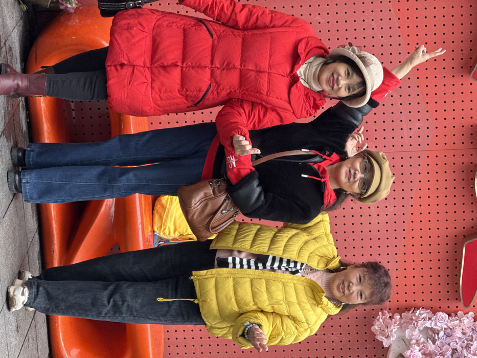 Three East Asian adult females are depicted standing and posing for a photograph outdoors in Liangjiang, China. They are positioned in front of a vibrant red wall featuring a uniform pattern of black circular perforations. Decorative elements on the wall include a partial light pink artificial floral arrangement on the upper right and a red high-heel shoe silhouette on the upper middle. A large, curvilinear orange bench-like structure is situated behind the subjects at waist level, with a yellow object resting on it. The ground is a wet, grey pavement.
The leftmost woman wears a beige woven hat, a long red quilted puffer coat over a light-colored turtleneck, black pants, and burgundy ankle boots. She smiles and holds hands with the middle woman. The middle woman wears a tan baker boy cap, glasses, a red hooded sweatshirt under a black faux fur jacket, blue jeans, and black shoes. She smiles, makes a 'V' sign with her raised right hand, holds hands with the leftmost woman, and has her left arm partially around the rightmost woman. The rightmost woman wears a light pink top, a black and white striped layer, a bright yellow quilted puffer jacket, dark blue/grey jeans, and white sneakers. She smiles, wears a beaded bracelet on her left wrist, and holds her left hand slightly open in a gesture. All three women maintain direct eye contact with the camera.