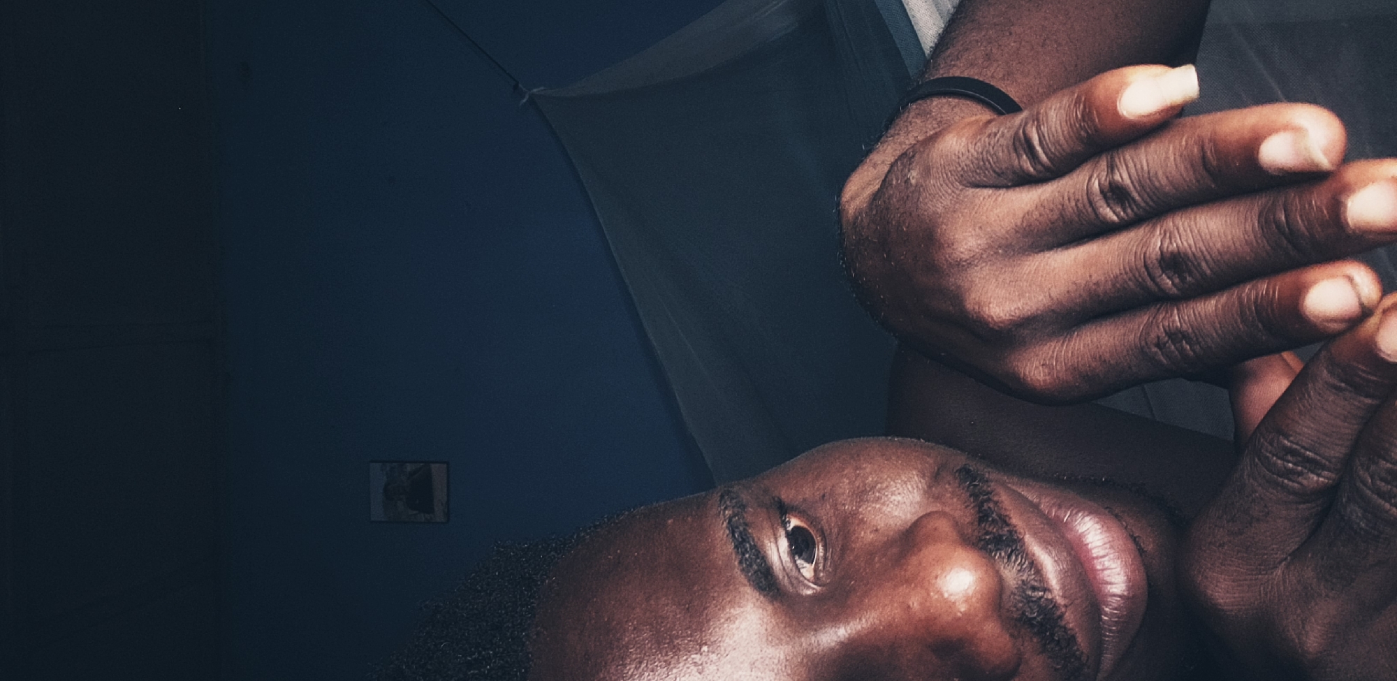 The image is a close-up, low-light, vertical shot of a dark-skinned man's face and hands. The man is lying down, with his head tilted to his left, and his gaze is directed downwards and slightly to the left. His left hand is resting on his face, with his fingers loosely touching his cheek. His right hand is positioned in the foreground, with his fingers extended and resting on a surface, possibly a bed. A black bracelet is visible on his right wrist. His skin is shiny, suggesting sweat or oil. A thin mustache is visible above his upper lip. In the background, a dark blue wall is visible with a small framed picture hanging on it. A white mosquito net is draped over something behind the man. The overall lighting is dim and moody, creating shadows and highlighting certain features. The scene appears to be indoors, likely in a bedroom, in Narayi, Nigeria.