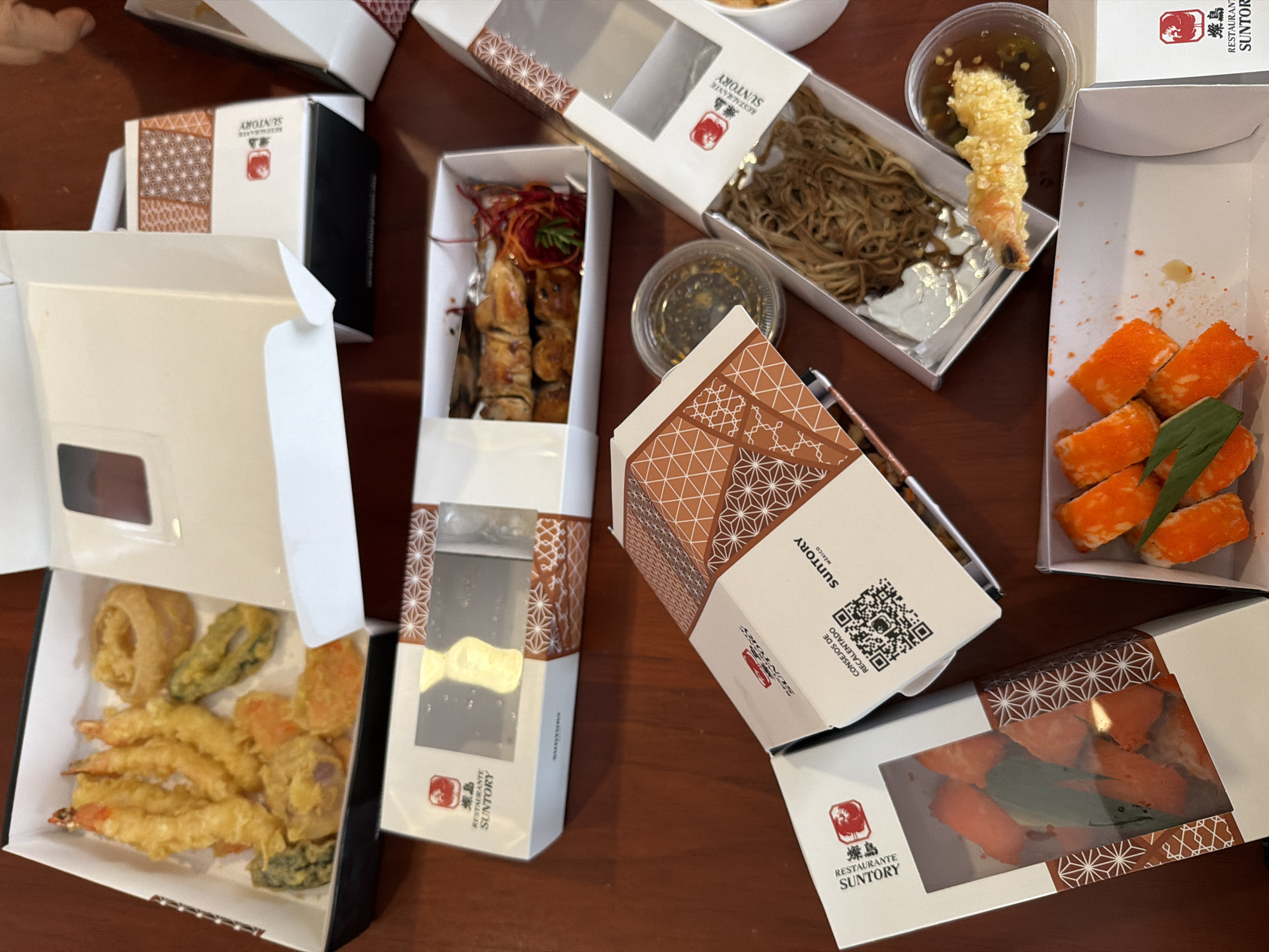 This is a photograph of various Japanese dishes from Restaurante Suntory in San Bernardino Tlaxcalancingo, Mexico, prepared for takeout. The dishes are contained in white cardboard boxes that bear the restaurant's logo and branding. The boxes are scattered on a dark wooden surface.
The image reveals the following:
- A box filled with tempura, including shrimp, vegetables, and perhaps squid.
- A box containing yakitori, garnished with shredded carrots.
- A box with soba noodles and tempura shrimp, accompanied by a dipping sauce in a clear plastic container.
- A box showcasing sushi rolls with orange roe topping.
- A box displaying salmon nigiri.
The restaurant's name and logo are prominently displayed on the boxes, along with information about reheating instructions and the location in Mexico.
