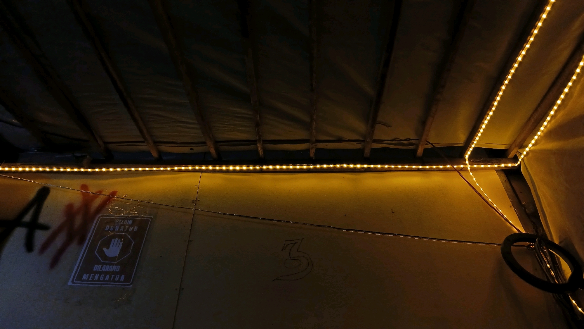 The image depicts the interior of a structure, likely a shed or makeshift room, with a canvas-like material stretched over wooden beams forming the ceiling. A string of yellow LED lights runs along the edge of the ceiling, casting a warm glow. The wall beneath the ceiling is a textured, yellowish surface, possibly concrete or painted wood.
On the wall, there is graffiti. On the left, a large, dark spray-painted letter "A" is visible, along with a reddish, smudged handprint or spray tag. To the right of this, a white sign with a dark border is affixed to the wall. The sign features a stylized white hand inside a hexagon, with text above and below it. The text above reads "SELAM DONATUR" and the text below reads "DILARANG MENGATUR". Translating this from Indonesian, the sign likely indicates "Do not disturb" or "No entry" for donors. Further to the right, a stylized number "3" is drawn or painted on the wall in black.
In the lower right corner, a black, circular object, possibly a tire or a ring, is hanging from a wire or chain. Electrical wires are also visible running across the wall. The overall impression is a dimly lit, utilitarian space with some evidence of unauthorized decoration. The location is identified as Adi Luhur, Indonesia.